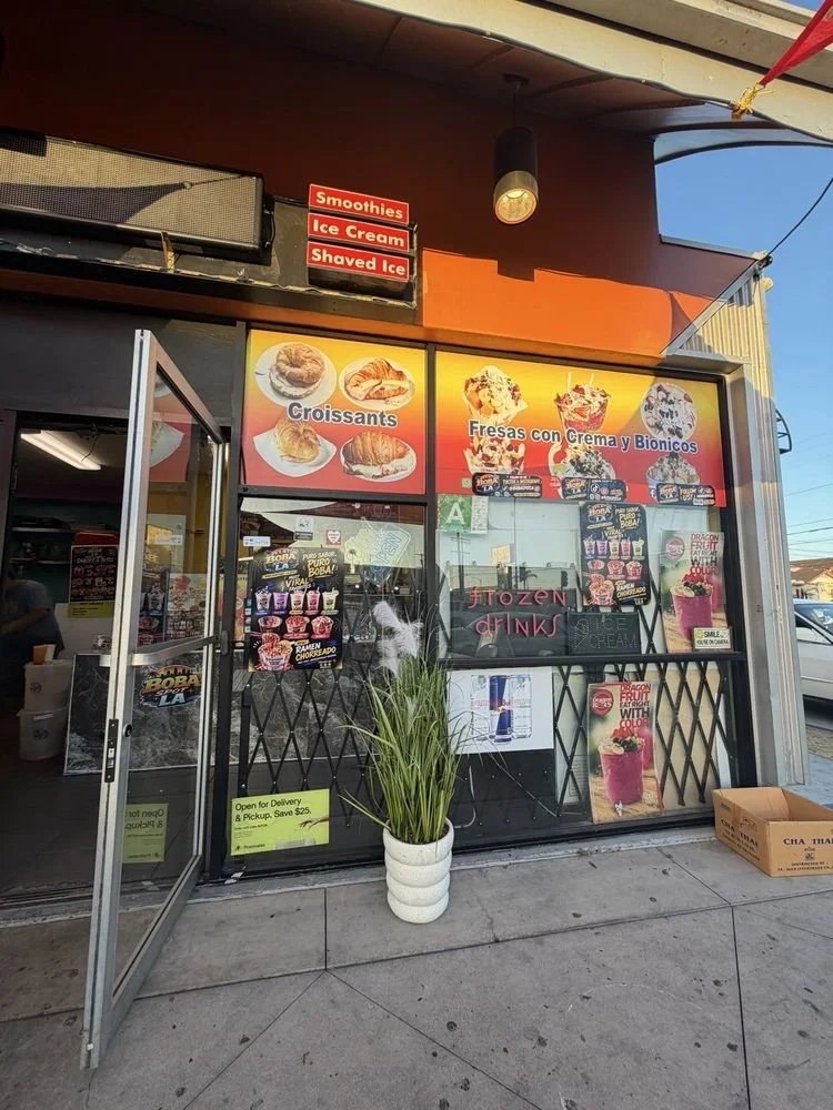 A storefront with signs advertising smoothies, ice cream, and shaved ice. Inside, a display with images of croissants and fruit, and a window with promotional posters for ice cream and frozen drinks. A potted plant is outside on the sidewalk.