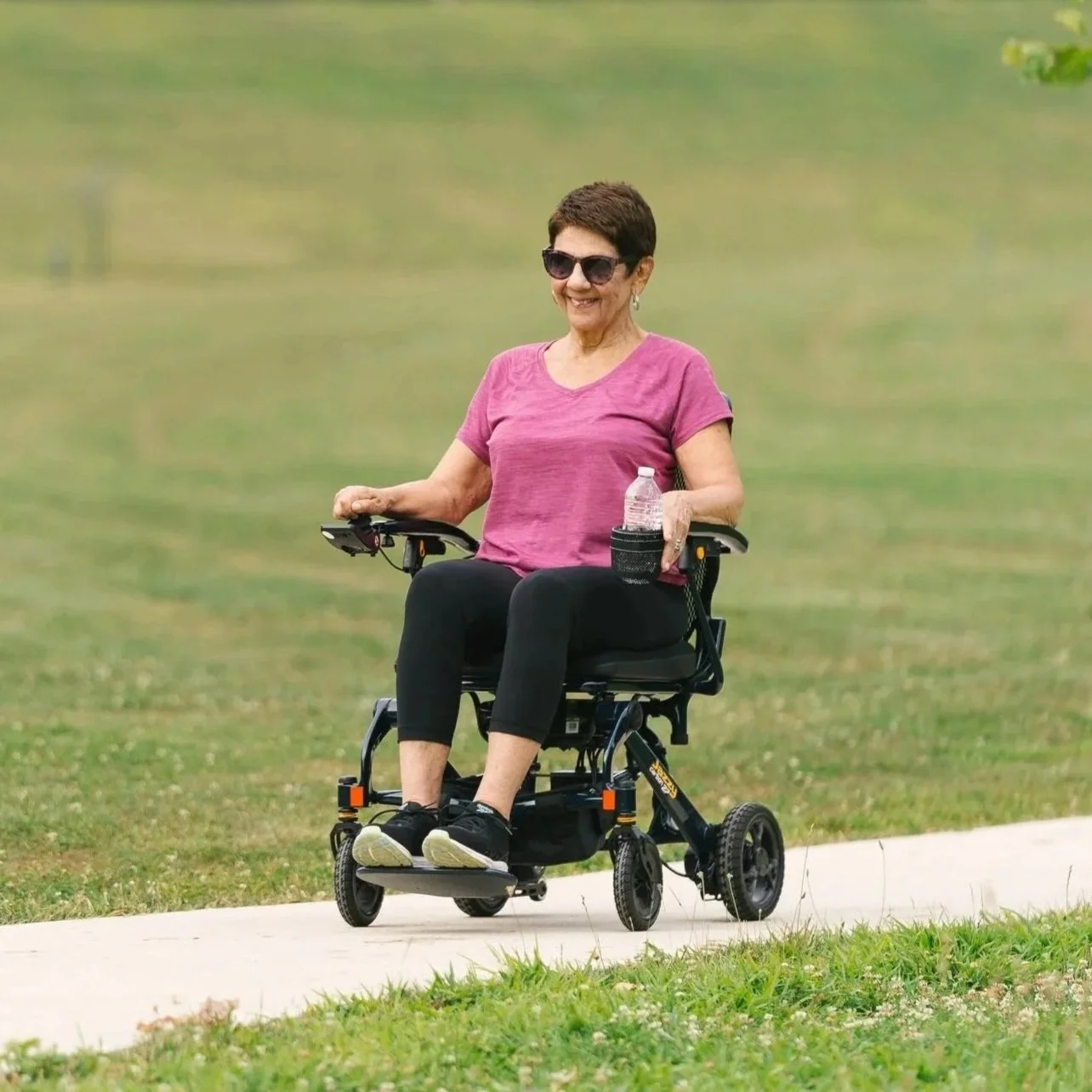 An elderly woman with short dark hair, wearing sunglasses, a pink T-shirt, black leggings, and black sneakers, riding a powered wheelchair outdoors on a paved path in a grassy park, holding a water bottle in a cup holder attached to the wheelchair.