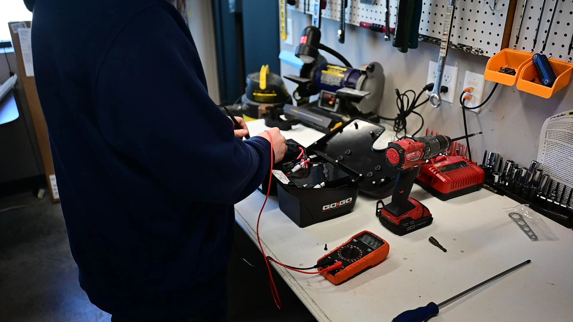 Person working on a battery or electronic device at a workbench with tools, drill, and multimeter in a workshop.
