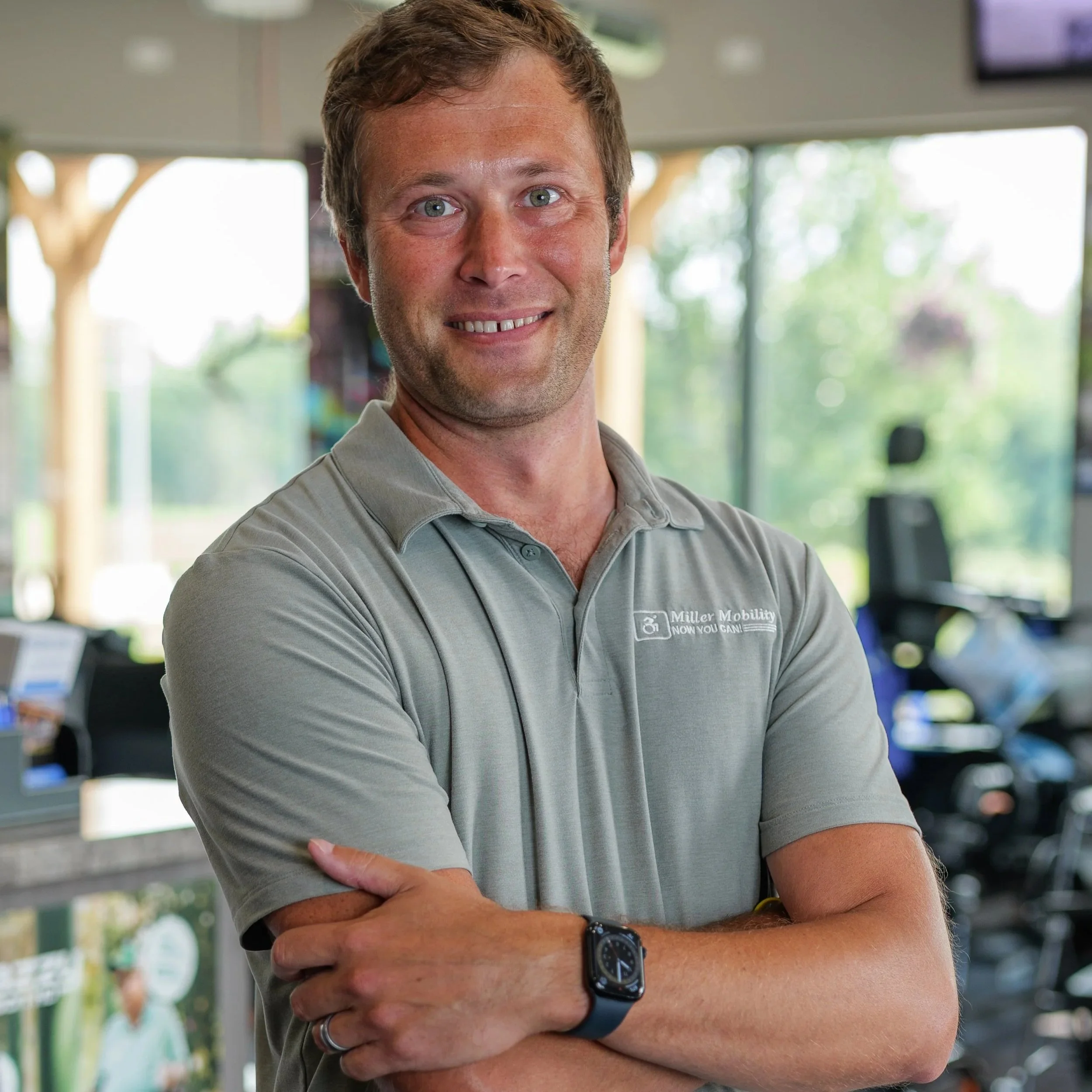 A man with light skin, short brown hair, and blue eyes smiling at the camera, wearing a gray polo shirt with a logo and text, standing indoors with large windows and greenery outside behind him.