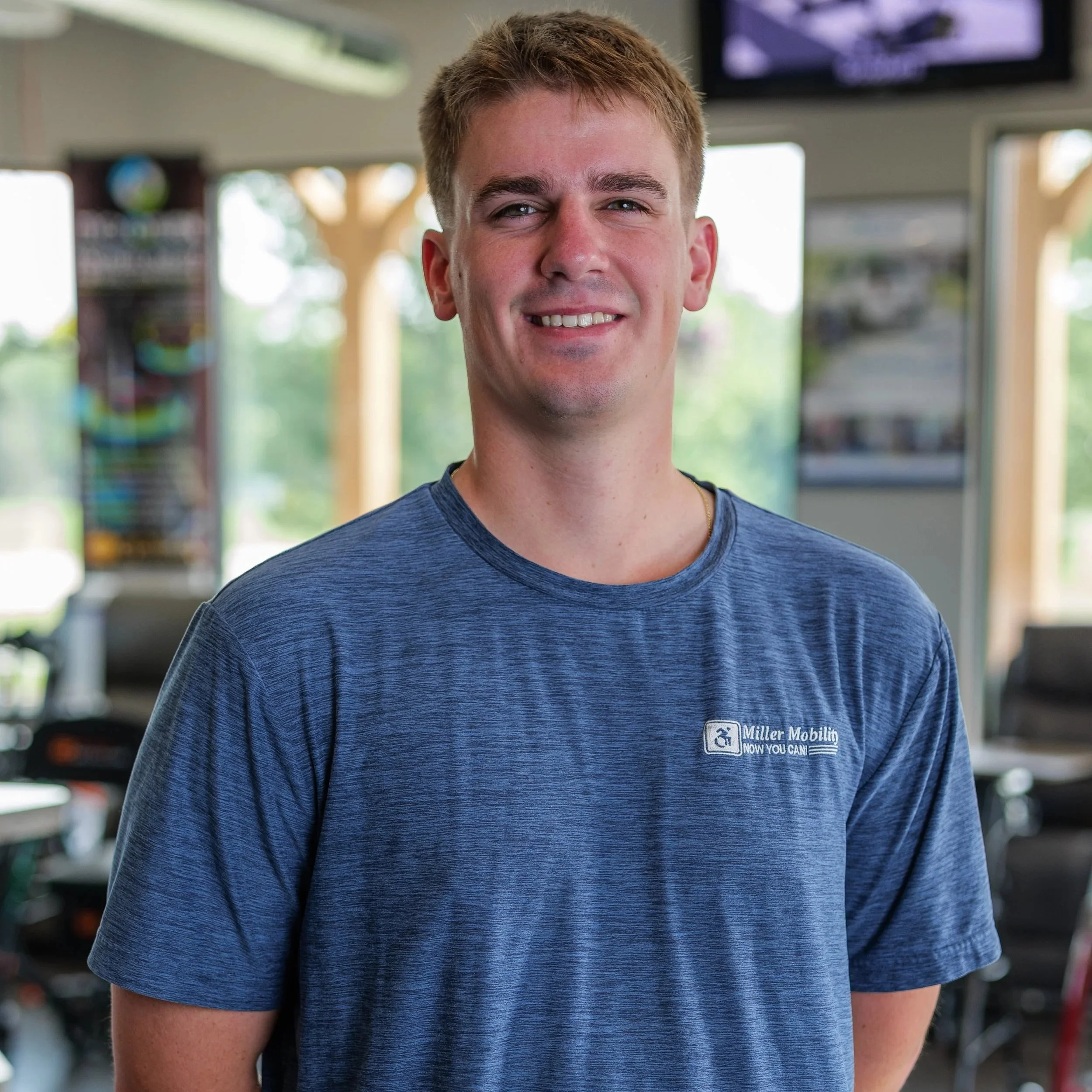 A young man wearing a blue t-shirt with a Miller Mobility Now You Can logo stands in a bright indoor space, smiling at the camera.