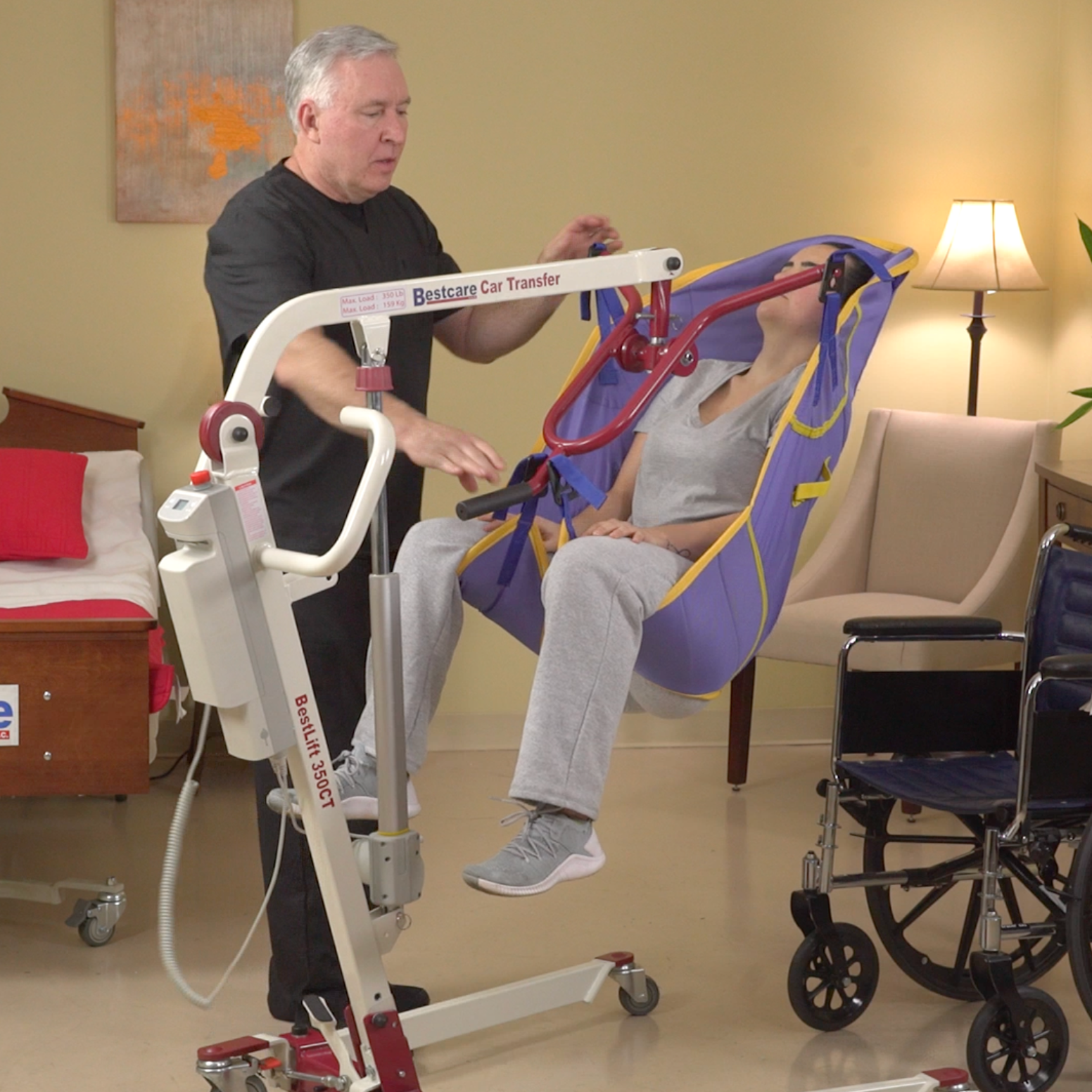 A man in black medical scrubs assisting a woman in a powered patient lift, with hospital chairs and a lamp in the background.