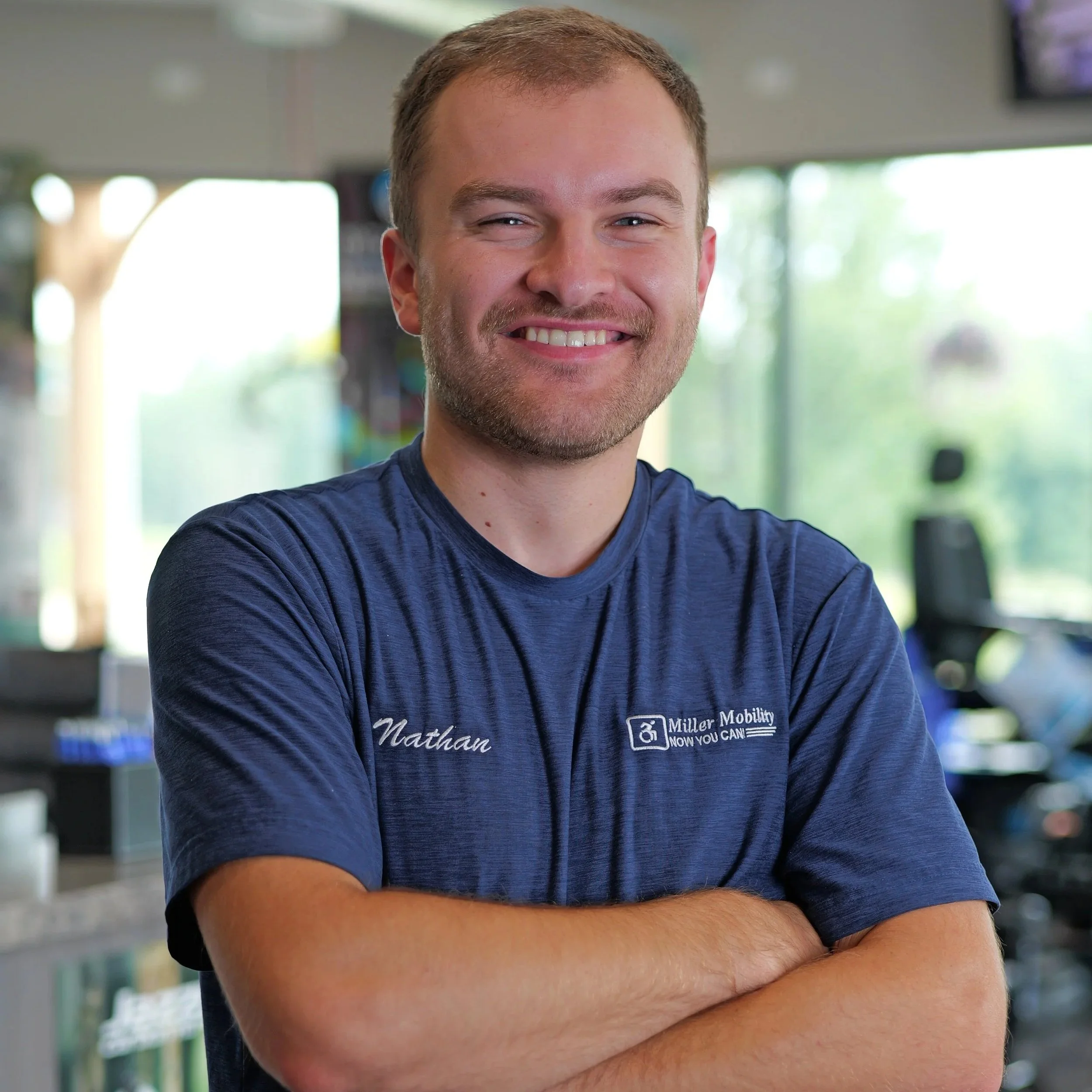 Close-up of a smiling young man with short light brown hair and a light beard, wearing a dark blue shirt with the name 'Nathan' and a logo on it, standing indoors with blurred background.