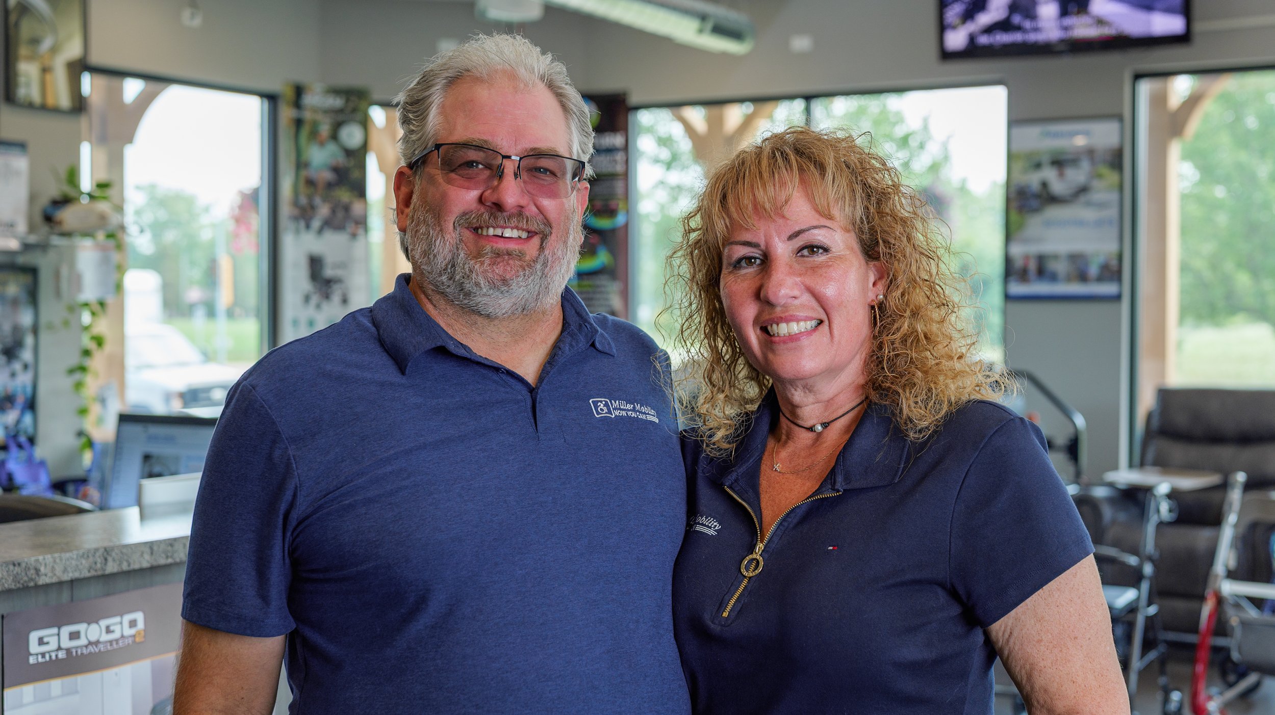 A smiling older man with gray hair, beard, and glasses standing beside a smiling woman with curly blonde hair, in an indoor setting with large windows and various equipment in the background.