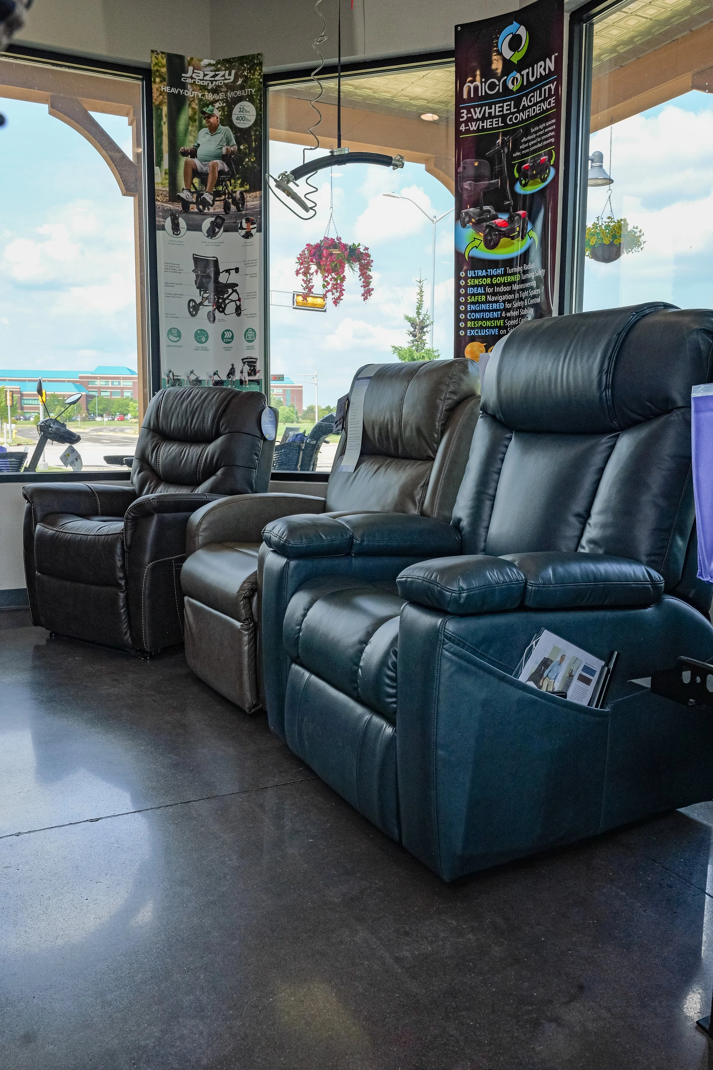 Indoor waiting area with black leather chairs, informational brochures, and promotional posters for mobility scooters on the walls, with a large window revealing a parking lot and blue sky outside.