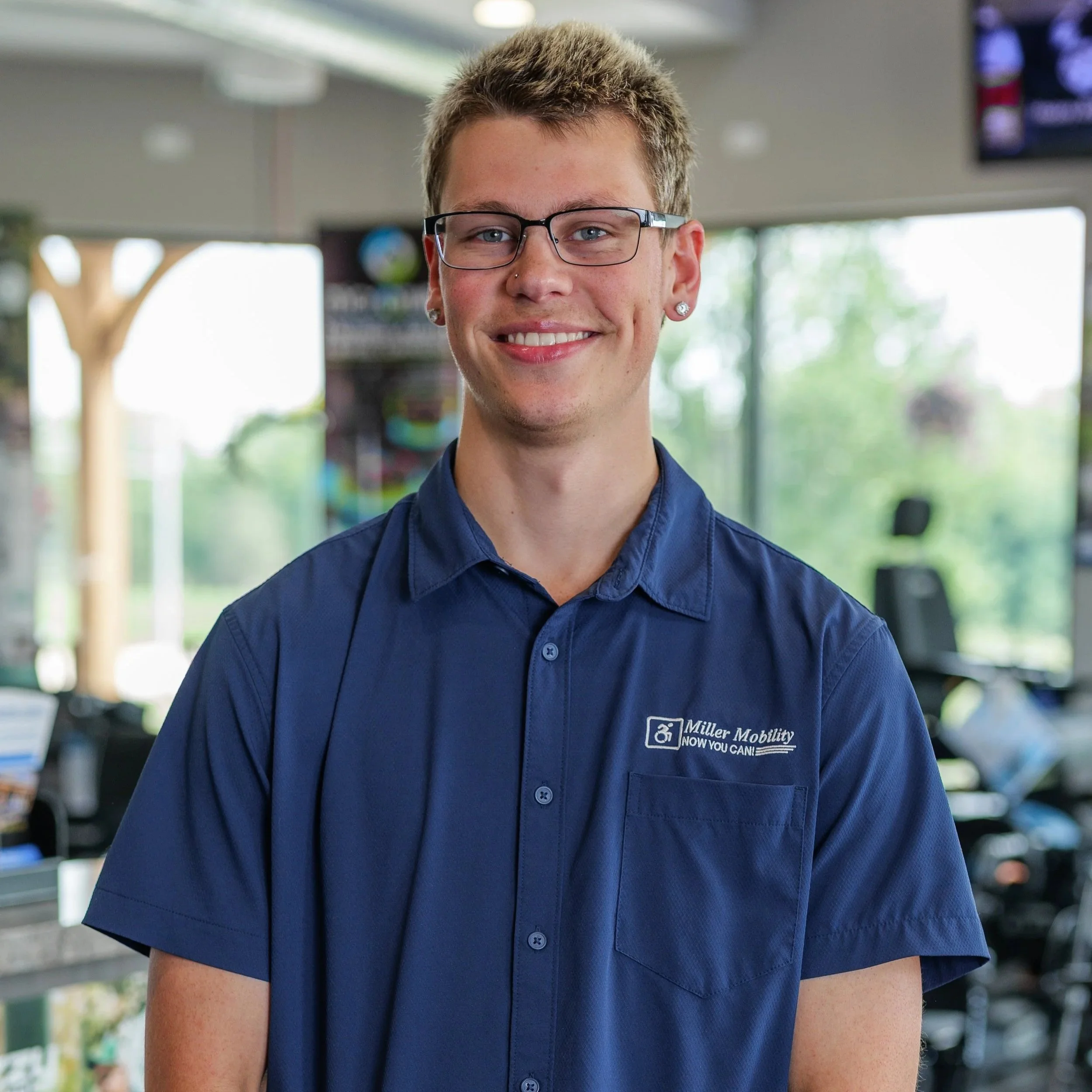 A young person with short, light brown hair, wearing glasses and a blue collared shirt with 'Miller Mobility' embroidered on it, smiling inside a store or office with shelves and a window in the background.