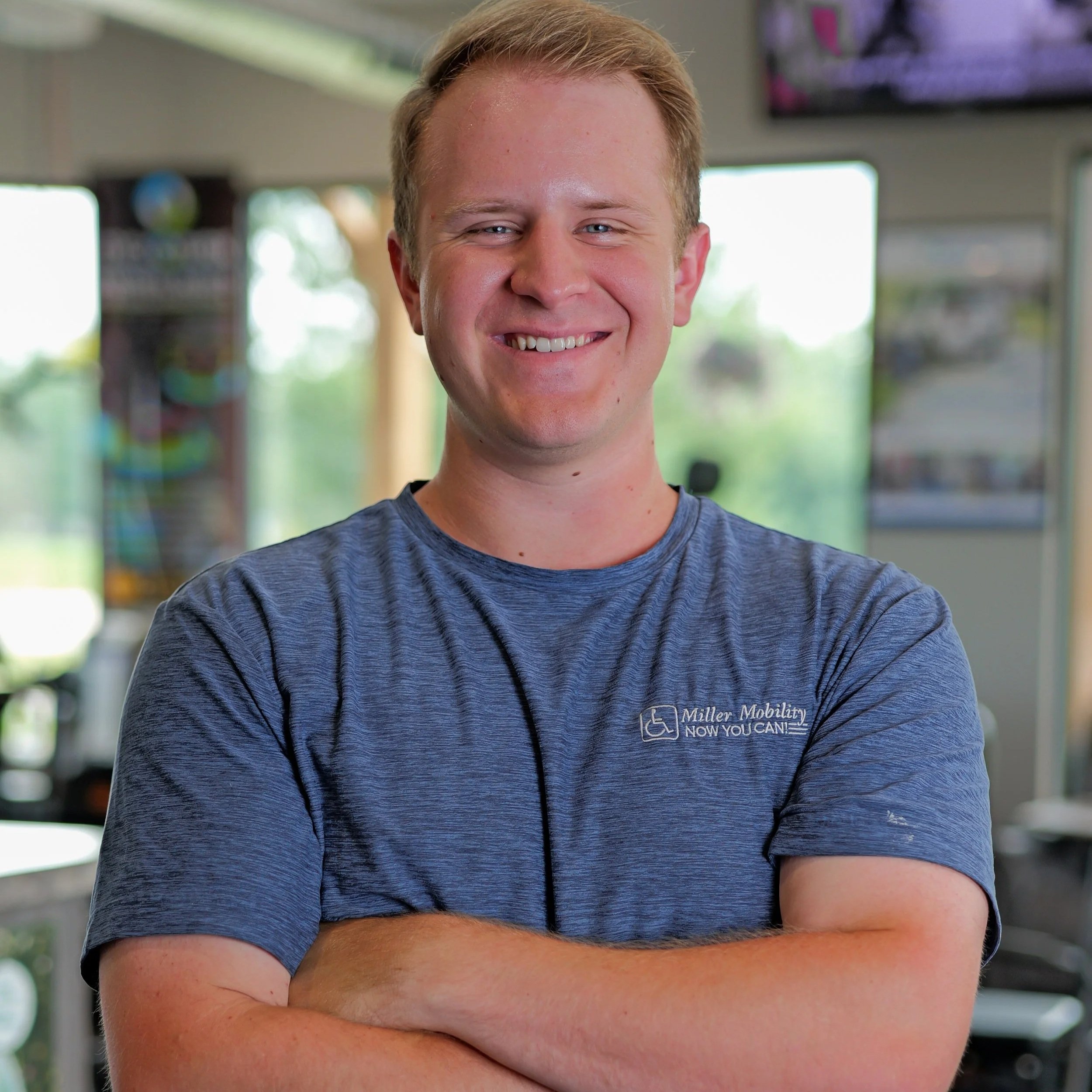 A young man with short blond hair smiling, wearing a gray T-shirt with a logo that promotes mobility for people with disabilities. He is standing indoors near large windows with blurred greenery outside.