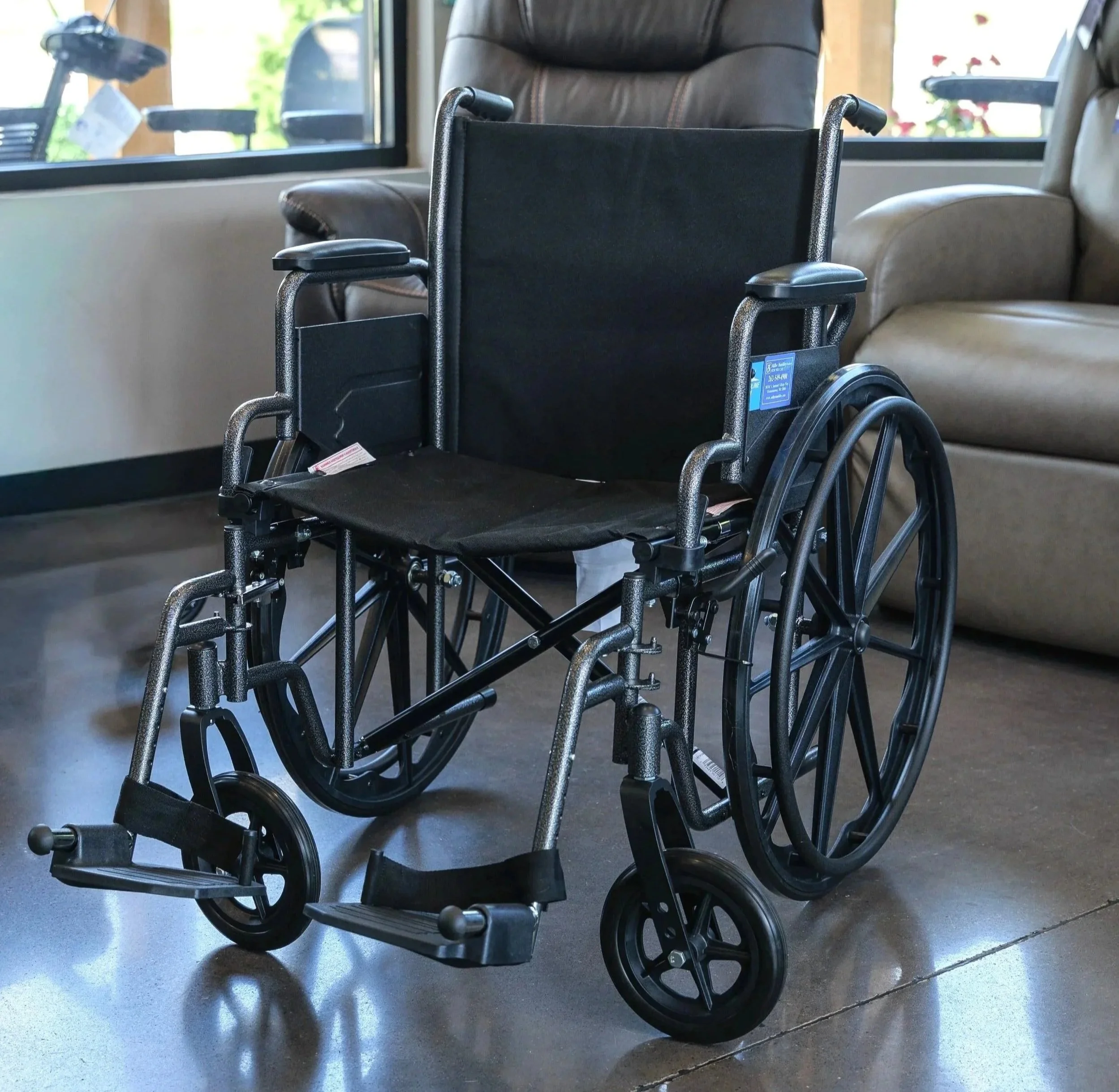 An empty wheelchair placed inside a room with large windows, a brown armchair, and sunlight streaming in from outside.