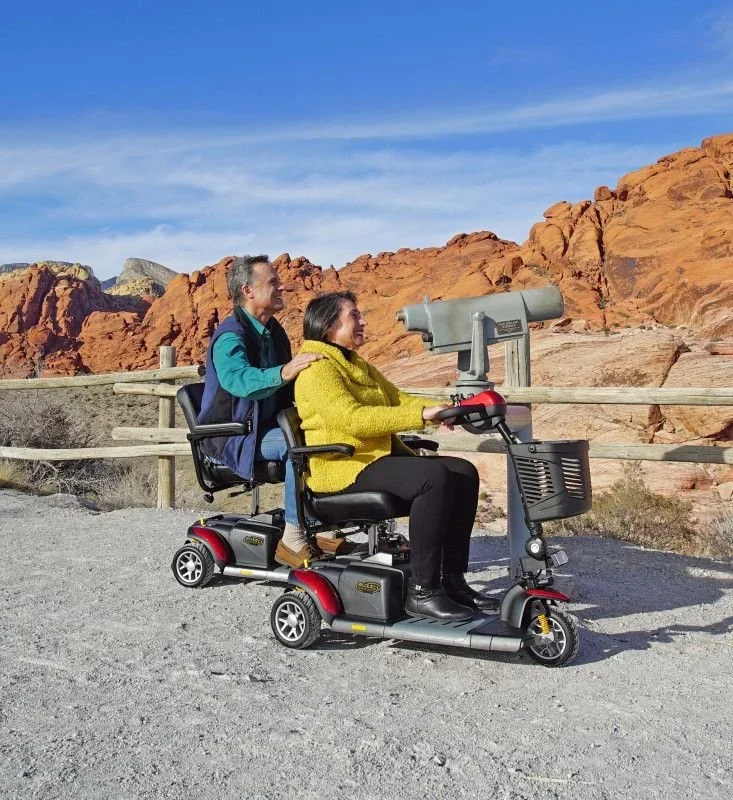 Two women riding a mobility scooter outdoors with a view of orange rocky cliffs and a blue sky in the background.