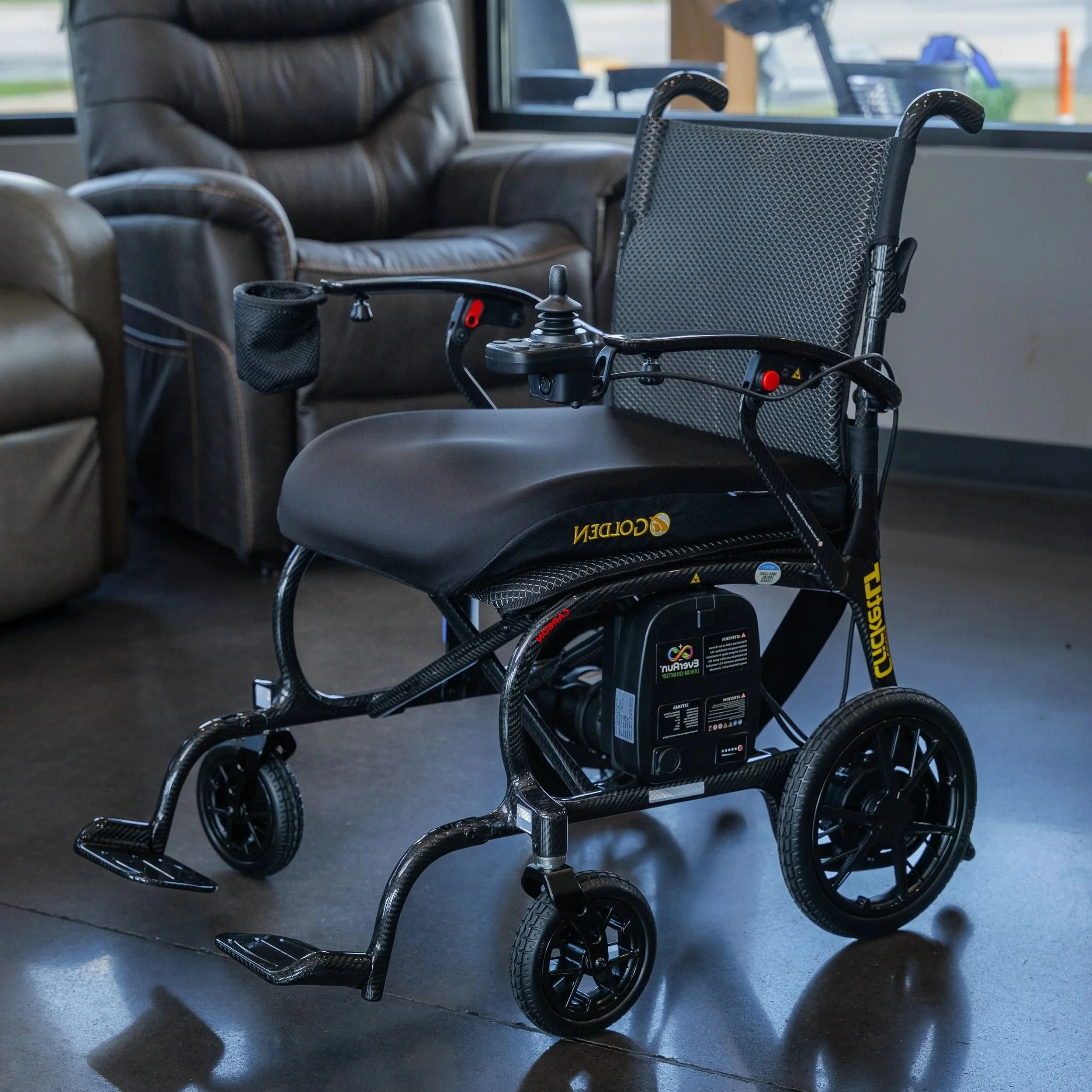 A power wheelchair with a black mesh backrest and black frame, positioned in front of brown leather recliner chairs in an indoor setting near a window with a view outside.