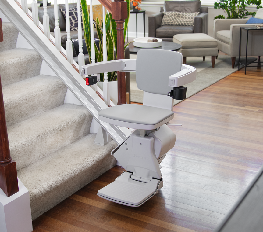 Stairlift attached to stairs in a living room with beige carpet, wooden handrail, and furniture including a gray sofa, beige ottoman, and plants.