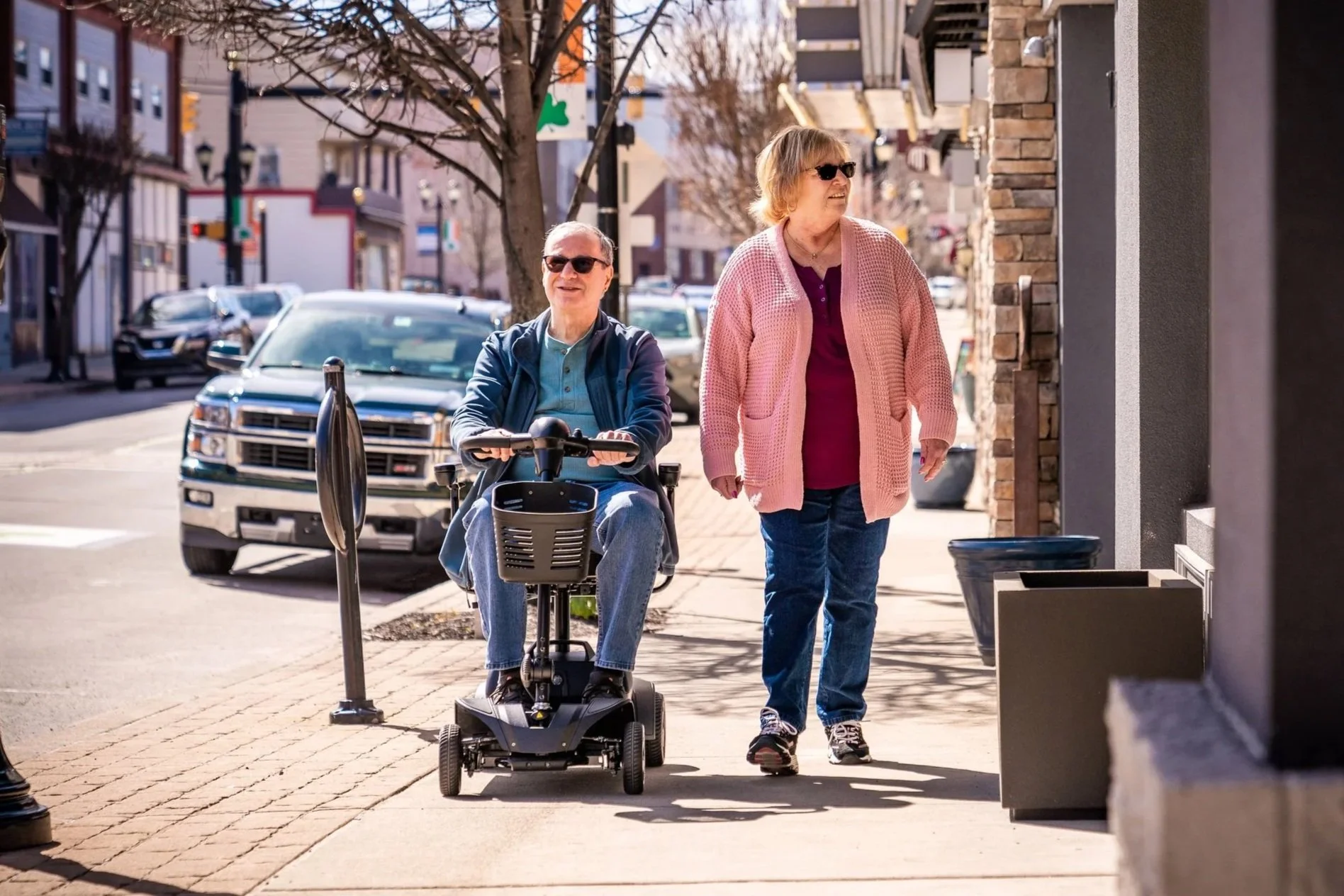 An elderly man using a mobility scooter and an elderly woman walking alongside on a city sidewalk during daytime. The man is smiling and wearing sunglasses, a blue jacket, and a light-colored shirt. The woman is wearing sunglasses, a pink cardigan, and a purple top. Cars are parked along the street, and there are buildings with storefronts, leafless trees, and a clear sky in the background.