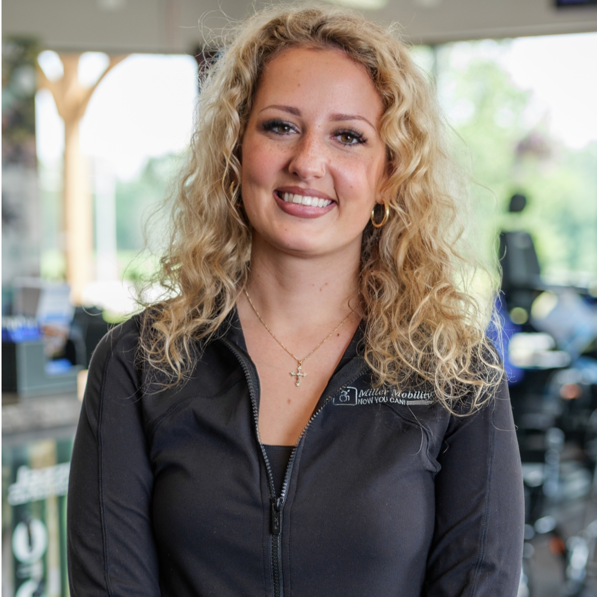 A woman with curly blonde hair smiling at the camera, wearing a black zip-up jacket with an embroidery logo, jewelry including a cross necklace and hoop earrings, in an indoor setting with gym equipment and large windows in the background.