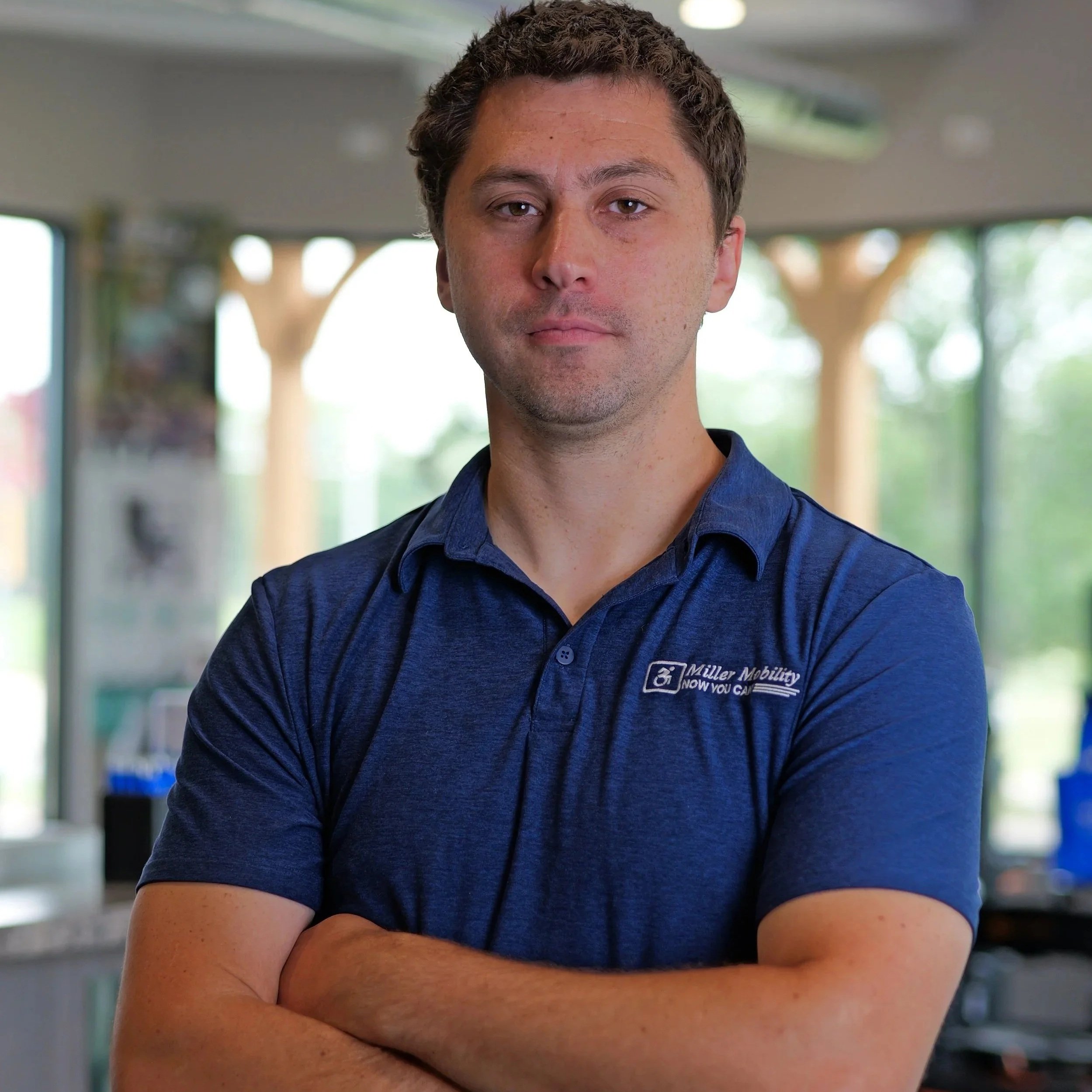 A man with short, dark curly hair and light skin wearing a dark blue collared shirt with a logo on it, standing indoors with a blurred background of windows and greenery outside.