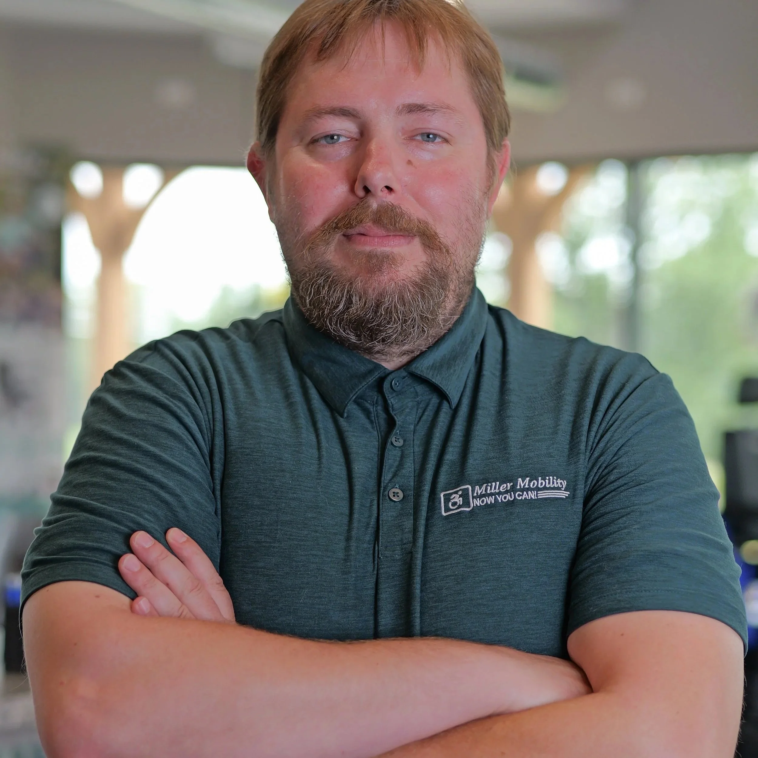 A man with red hair, a beard, and blue eyes, wearing a dark shirt with a Miller Mobility logo, standing indoors with blurred background, crossing his arms.