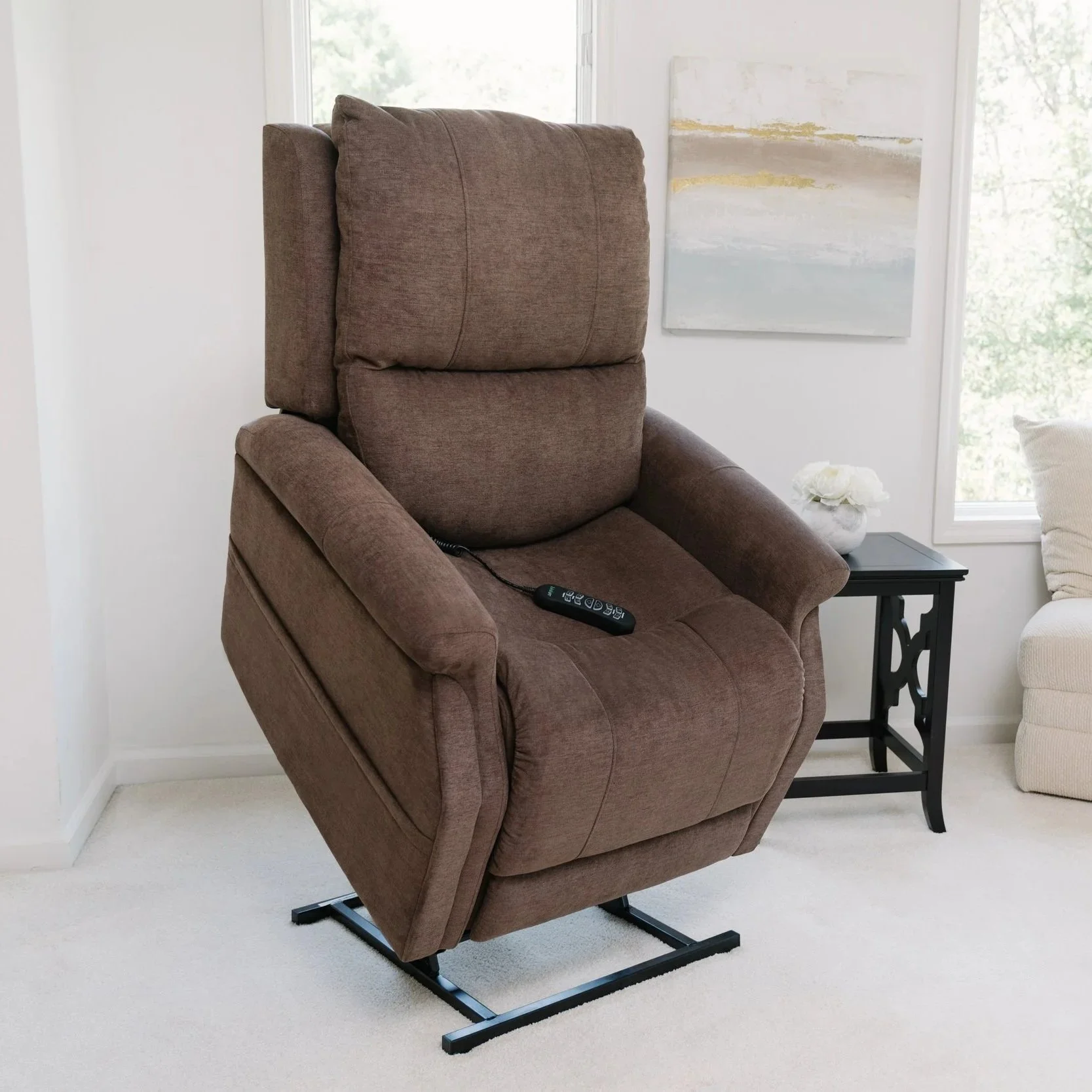 A brown upholstered recliner chair with a remote control resting on it, situated in a bright room with large windows, white walls, a side table with a white flower arrangement, and artwork hanging on the wall.