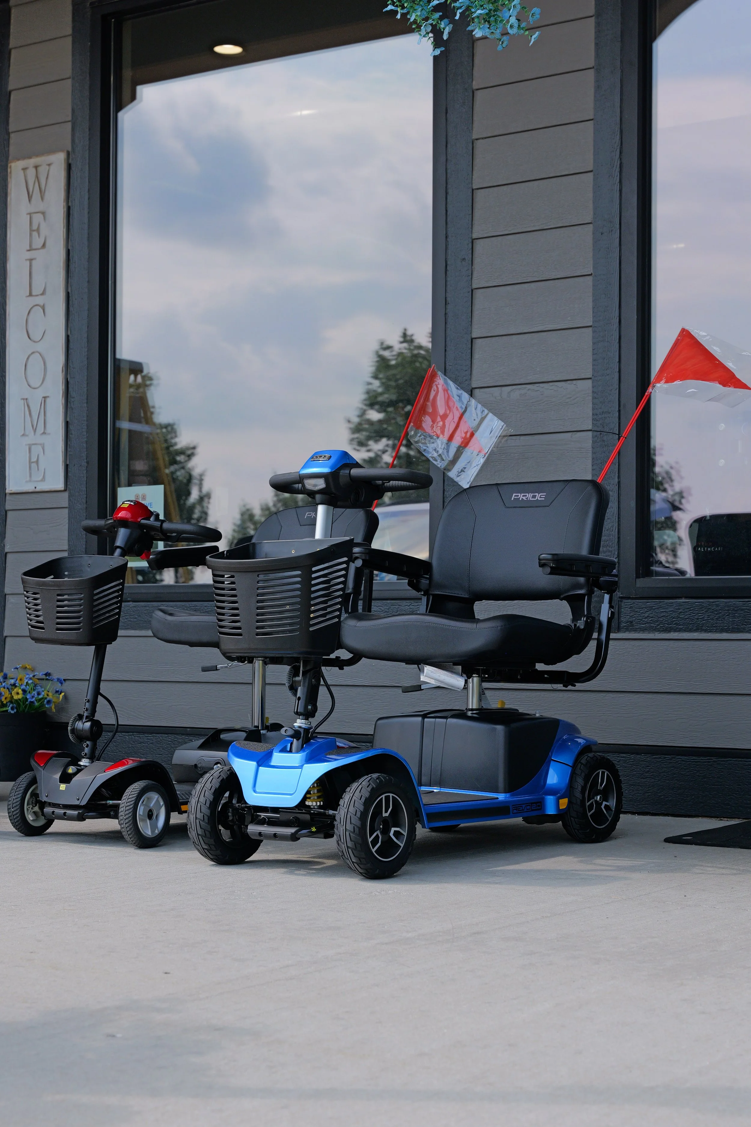 Outdoor scene with mobility scooters and a child's power ride-on vehicle parked in front of a building with a large window. The scene includes flags on the scooters and a welcome sign on the wall.
