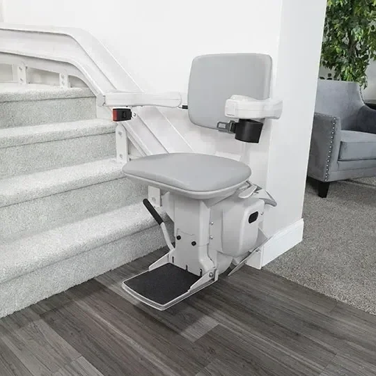 A stair lift with a gray seat and armrests installed at the bottom of carpeted stairs in a residential setting.