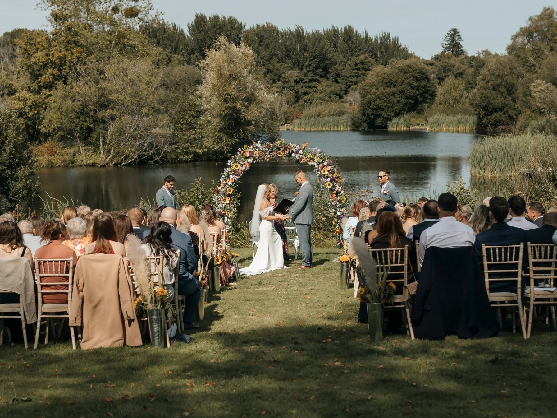 A wedding ceremony outdoors by a lake, with guests seated facing a couple exchanging vows under a floral arch, surrounded by trees with green and autumn foliage.