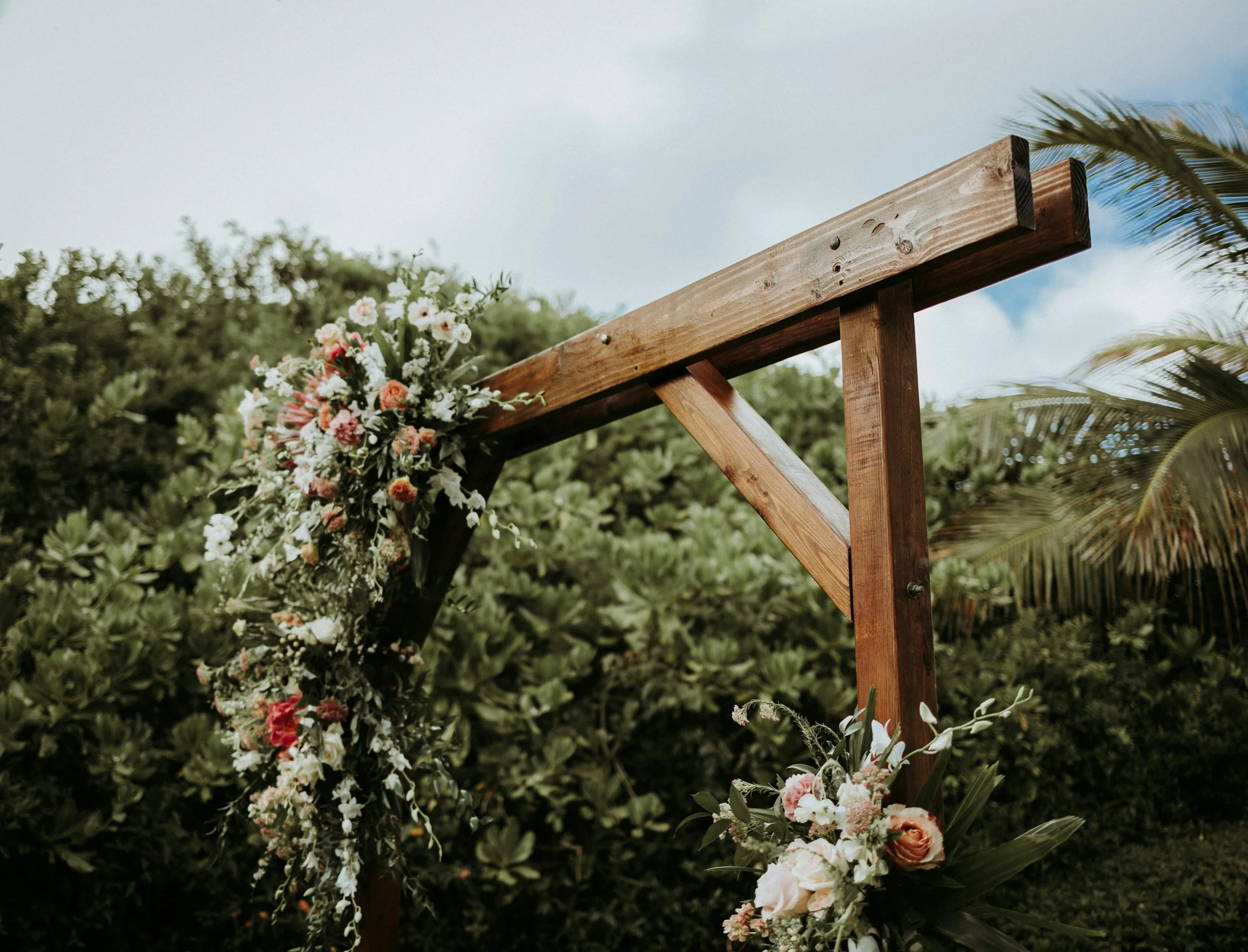 An outdoor wedding arch decorated with pink and white flowers, set against a backdrop of green bushes and a partly cloudy sky.