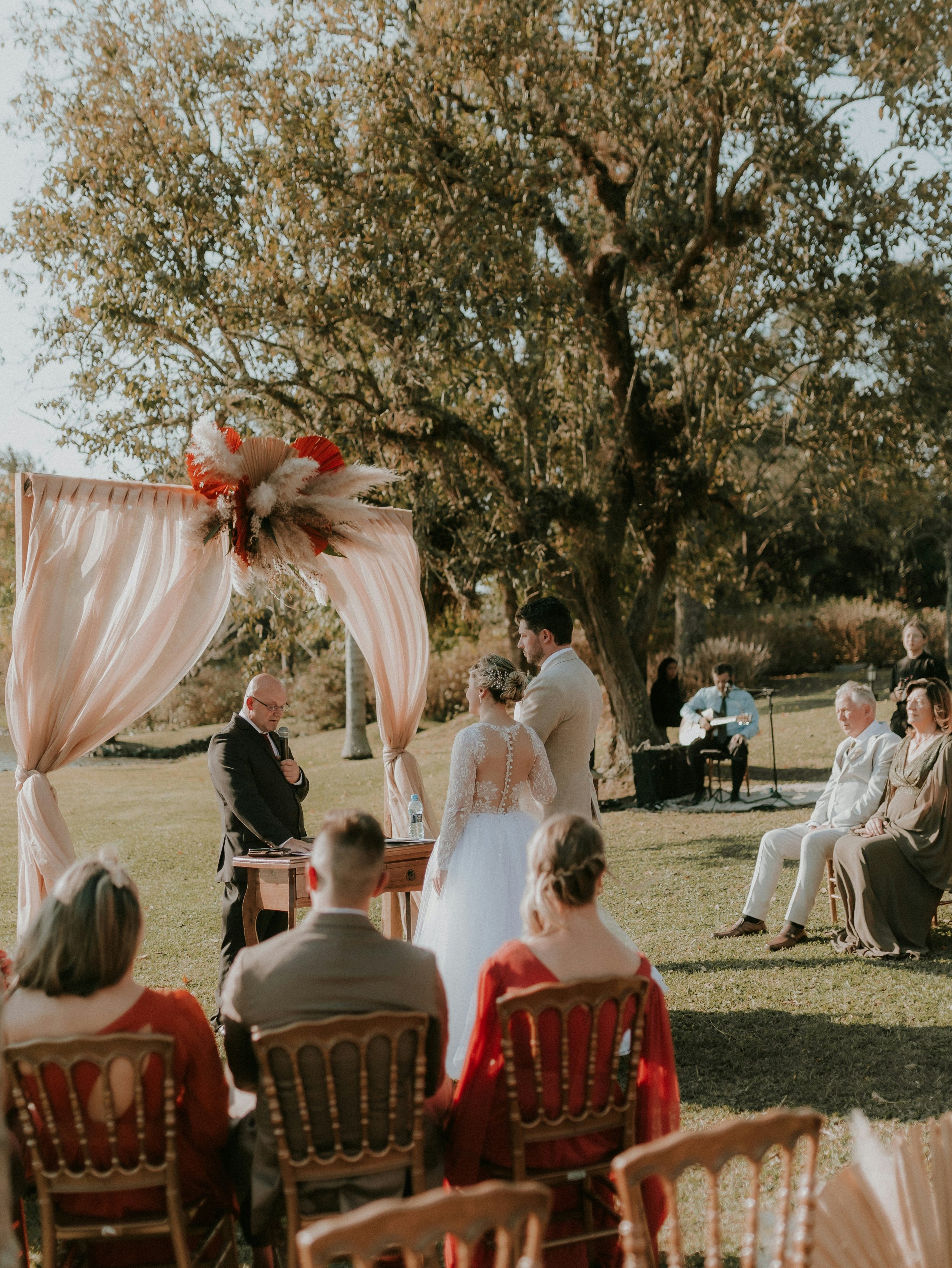 Outdoor wedding ceremony with bride and groom standing under a decorated arch, surrounded by seated guests and live musicians, with large trees and clear sky in the background.