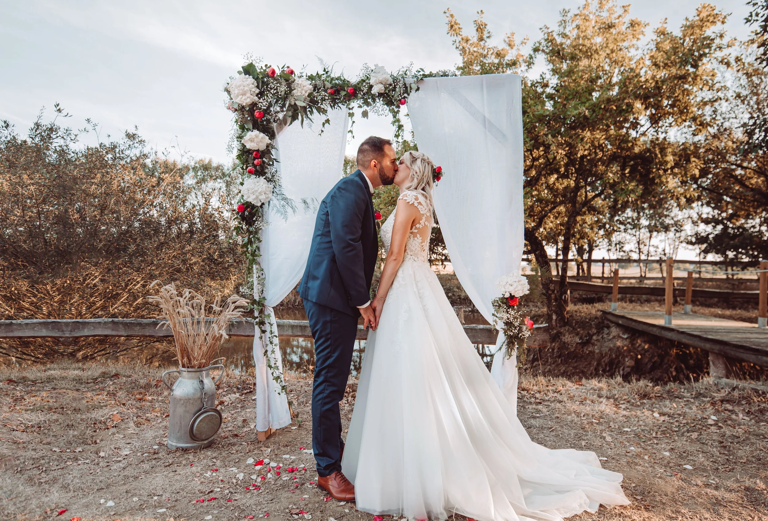 A couple dressed in wedding attire sharing a kiss under a decorated wedding arch outdoors, with trees and a cloudy sky in the background.
