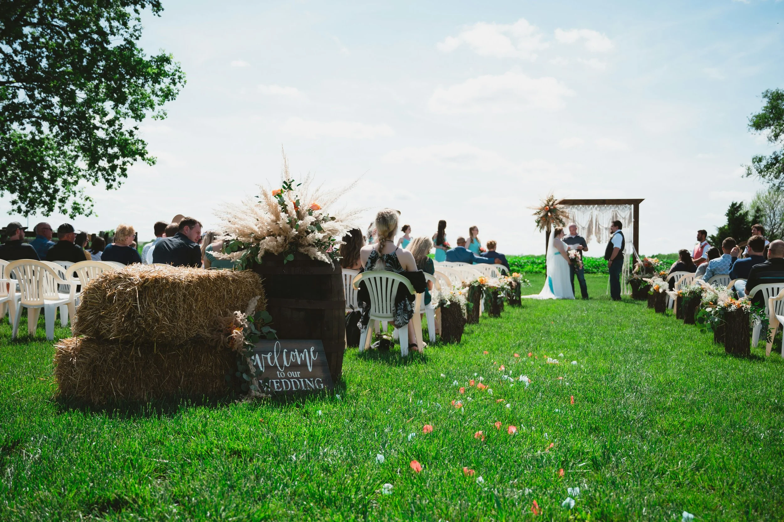 Outdoor wedding ceremony setup on a grassy field with chairs, floral arrangements, and an arch, under a partly cloudy sky, with trees and hay bales in the background.