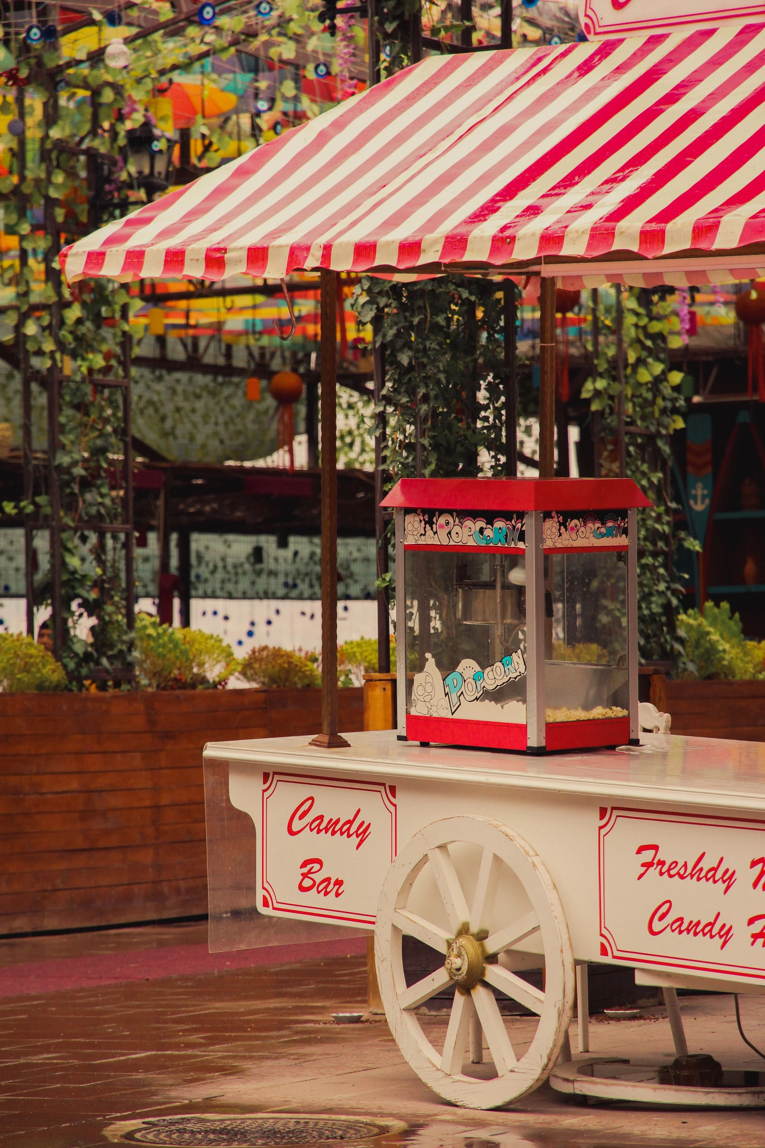 A red and white striped candy stand under a colorful outdoor canopy with an amusement park in the background.
