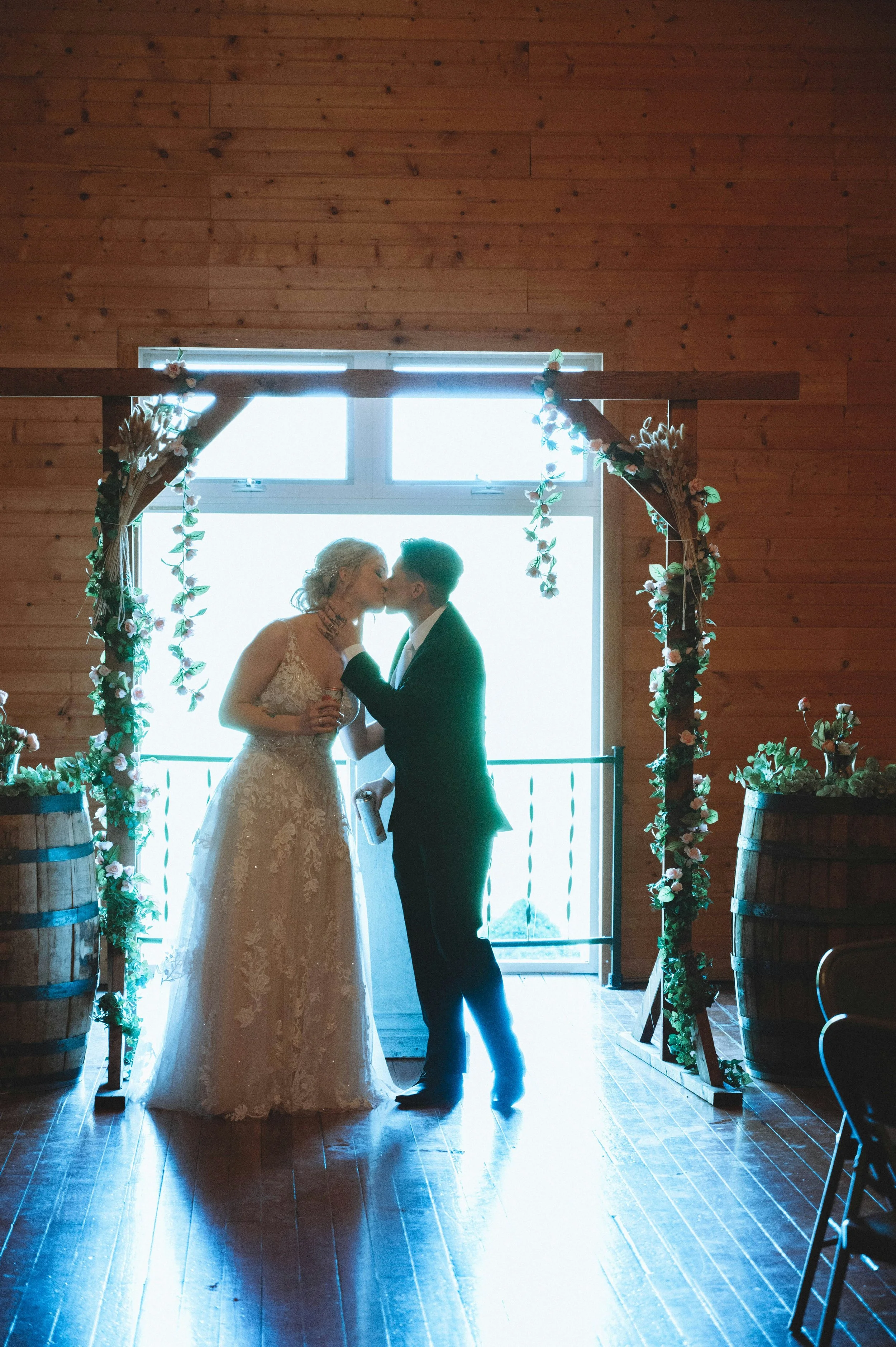 A bride and groom sharing a kiss under a floral arch inside a wooden venue with large windows and wooden barrels.