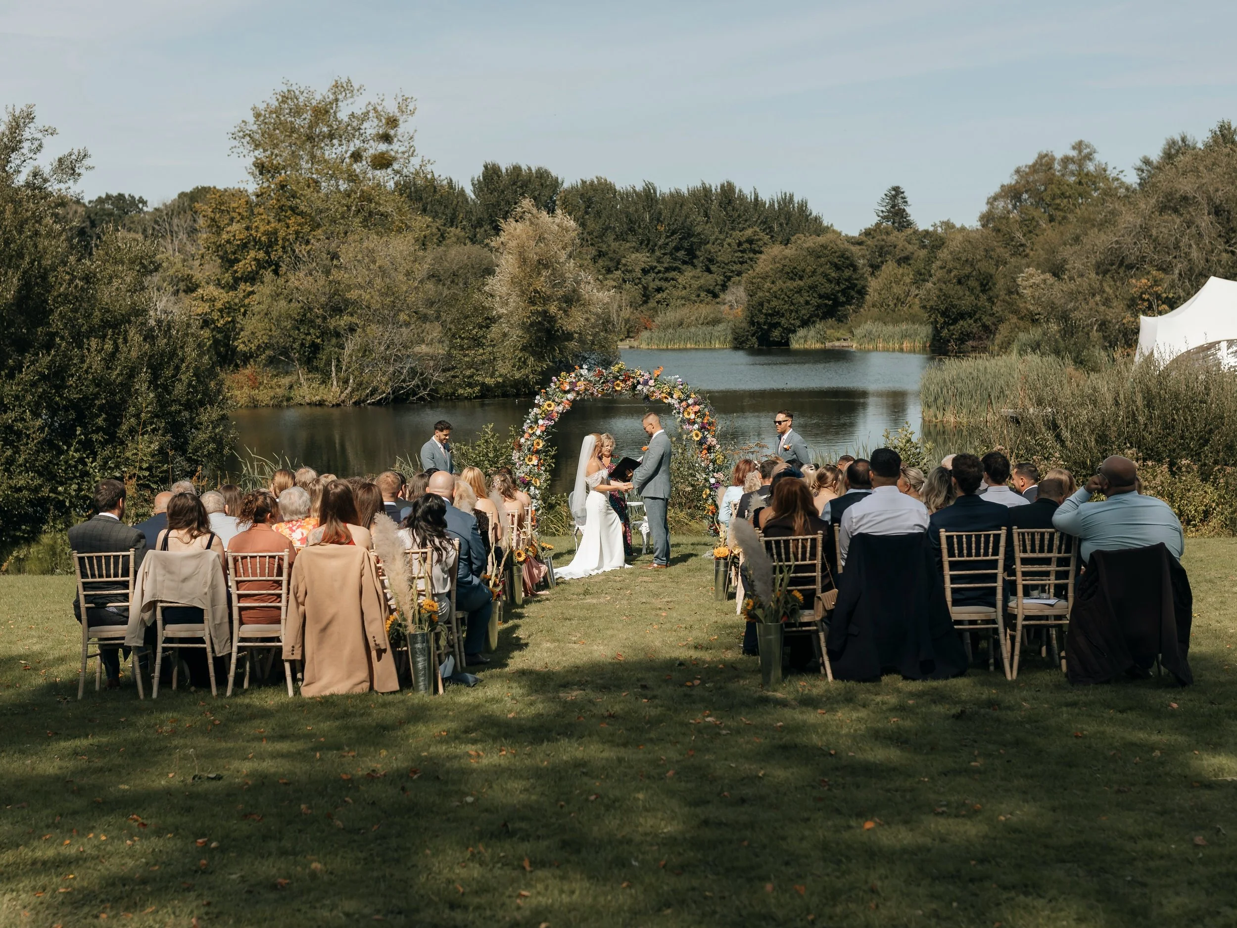 An outdoor wedding ceremony by a lake with guests seated, a bride and groom exchanging vows under a floral arch, surrounded by trees and a white tent in the background.