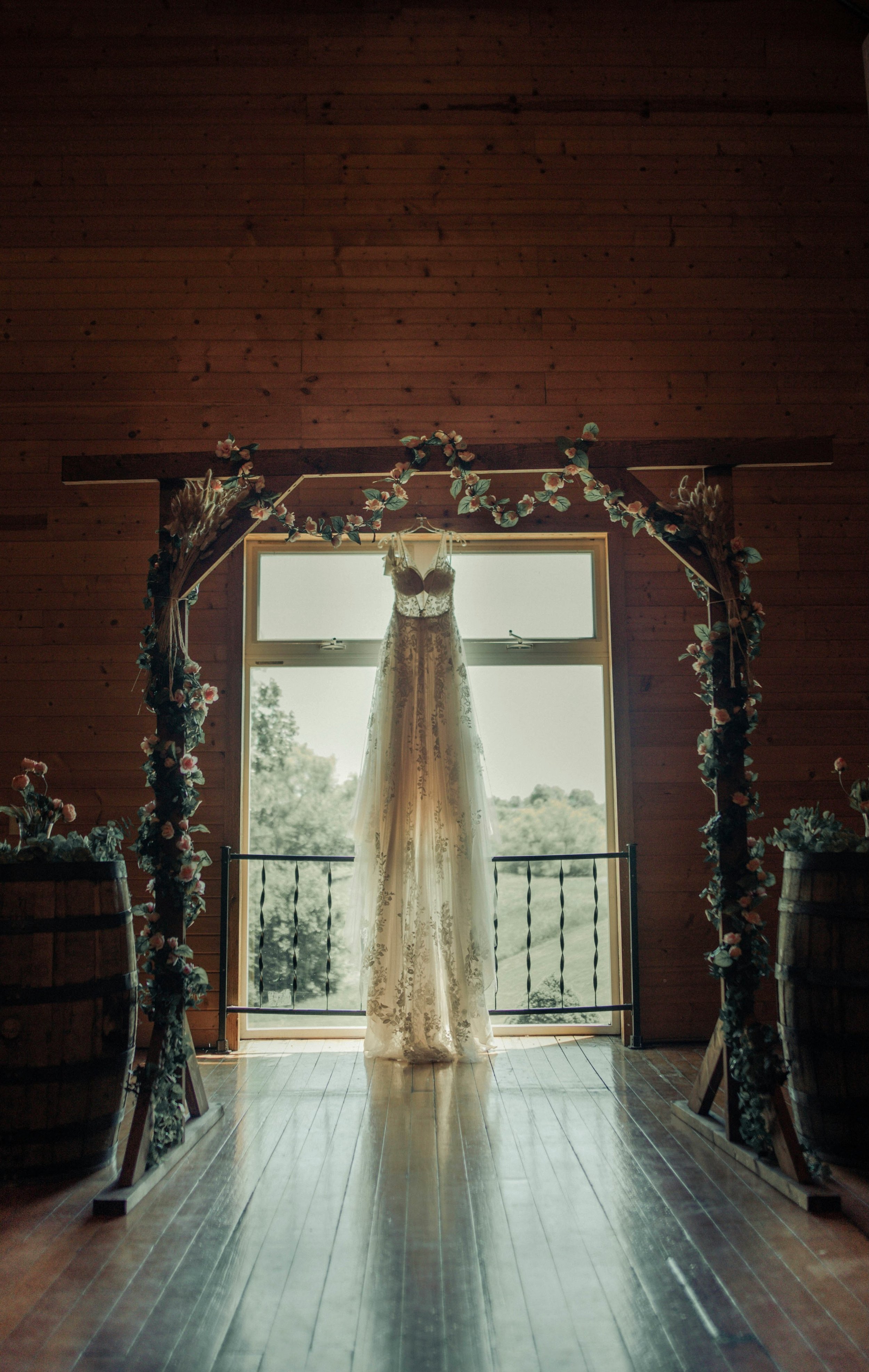 A wedding dress hangs on a wedding arch in front of a large window in a wooden room decorated with floral garlands and barrels, with sunlight streaming in and green trees visible outside.