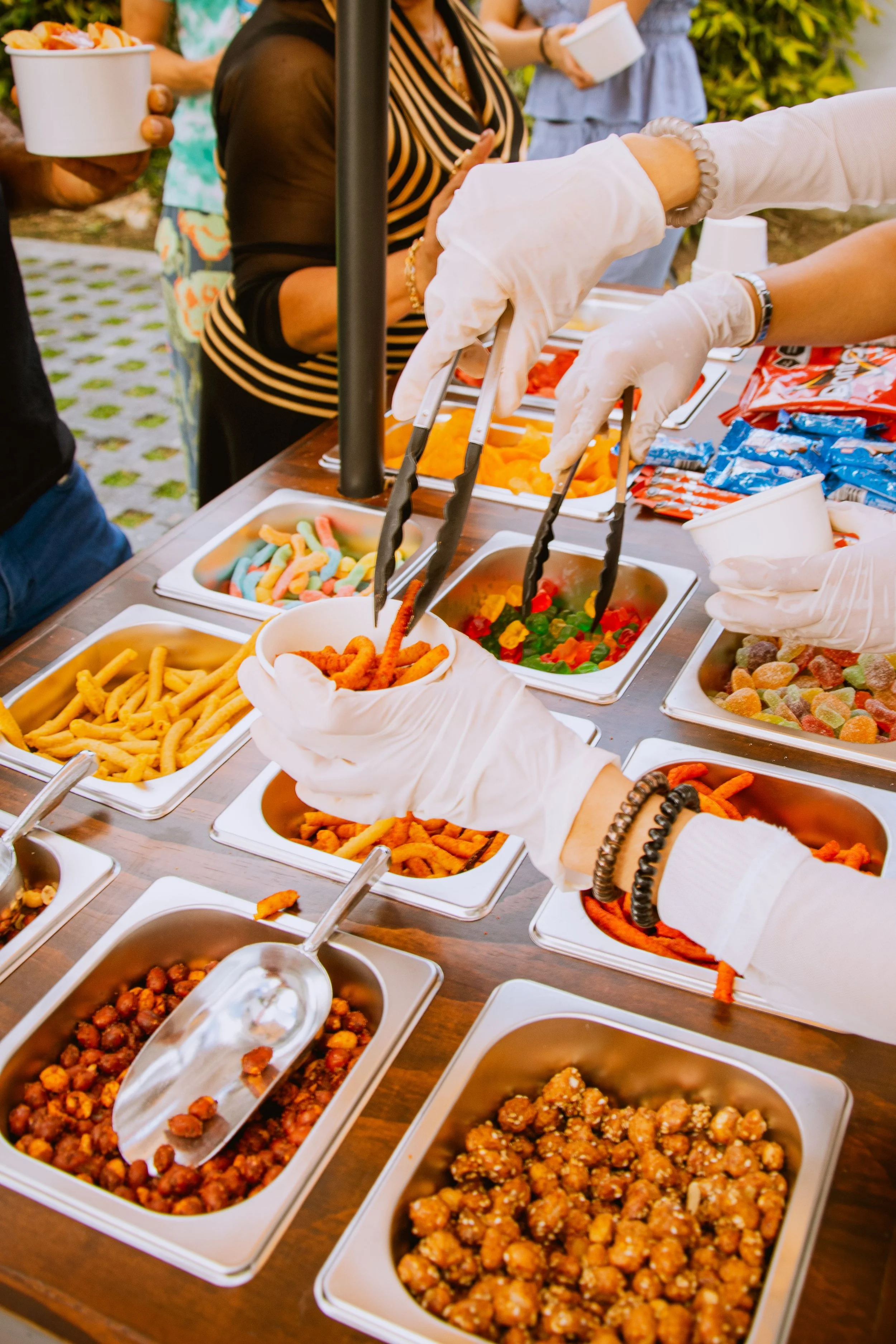 People serving themselves a variety of candy at a candy cart, including gummy candies, trail mix, and licorice, with some guests holding bowls and tongs.