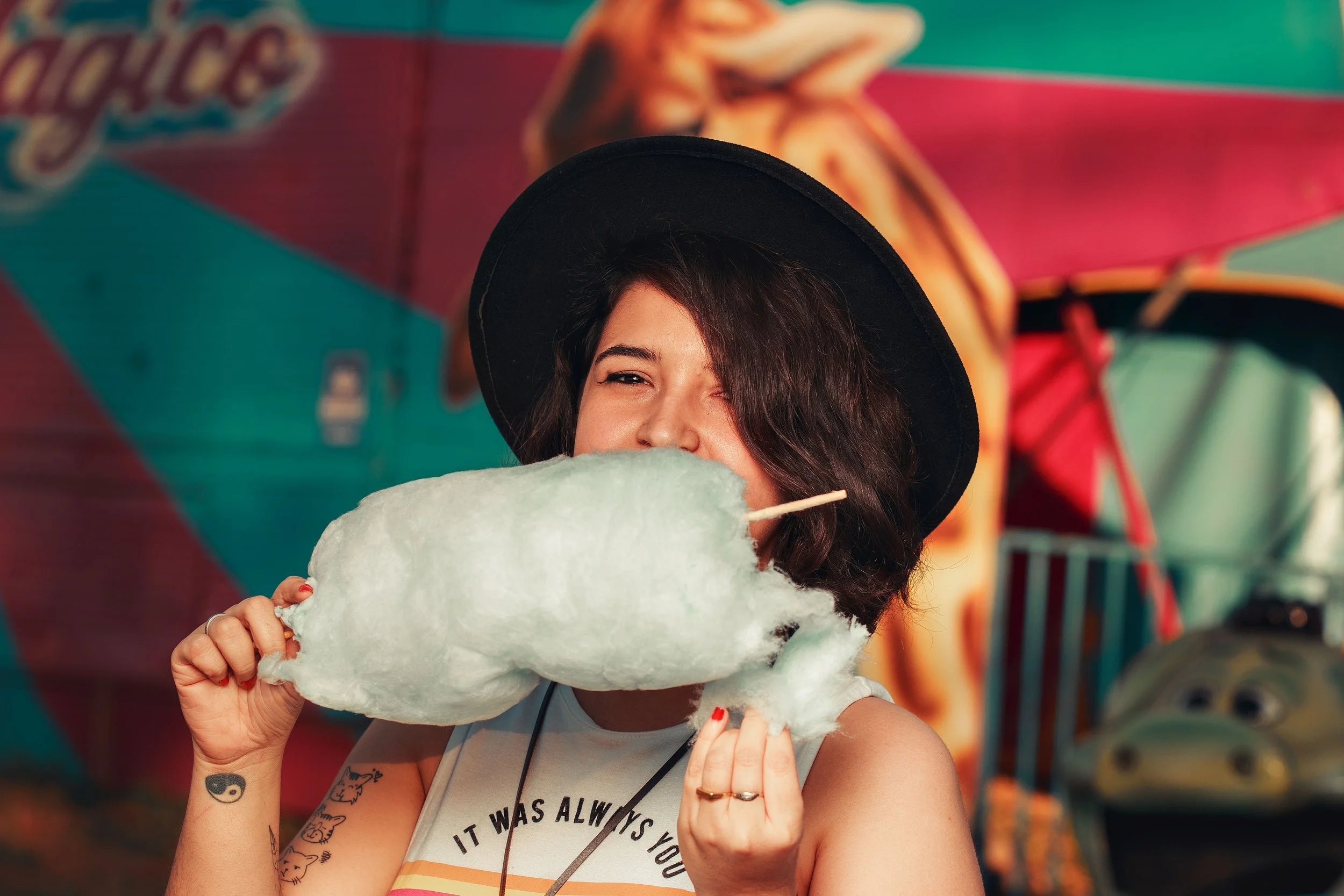 A woman with dark hair wearing a black hat and a sleeveless top smiling while holding cotton candy