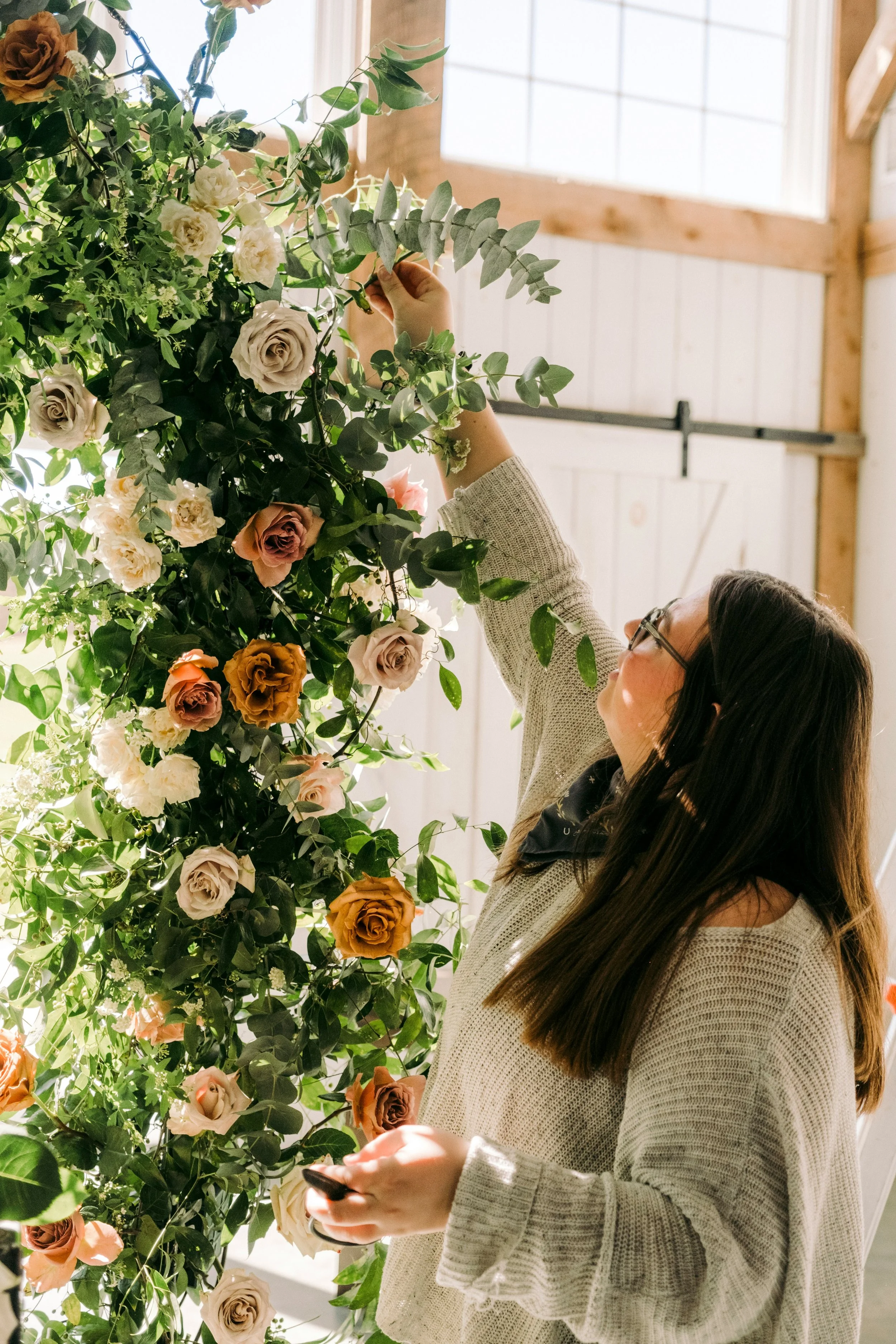 A woman with long brown hair and glasses is adjusting a large arrangement of various roses and greenery inside a barn-like space with wooden beams and a large window.