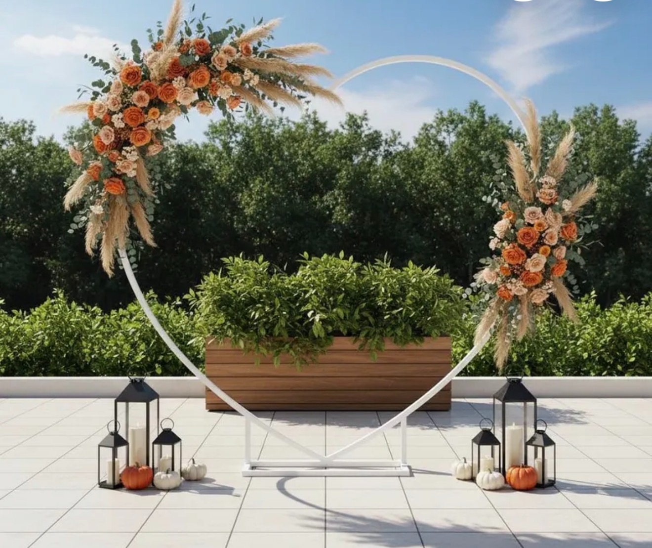 A decorative heart-shaped floral arch with peach, white, and blush flowers and pampas grass, surrounded by lanterns and pumpkins on a terrace with greenery in the background.