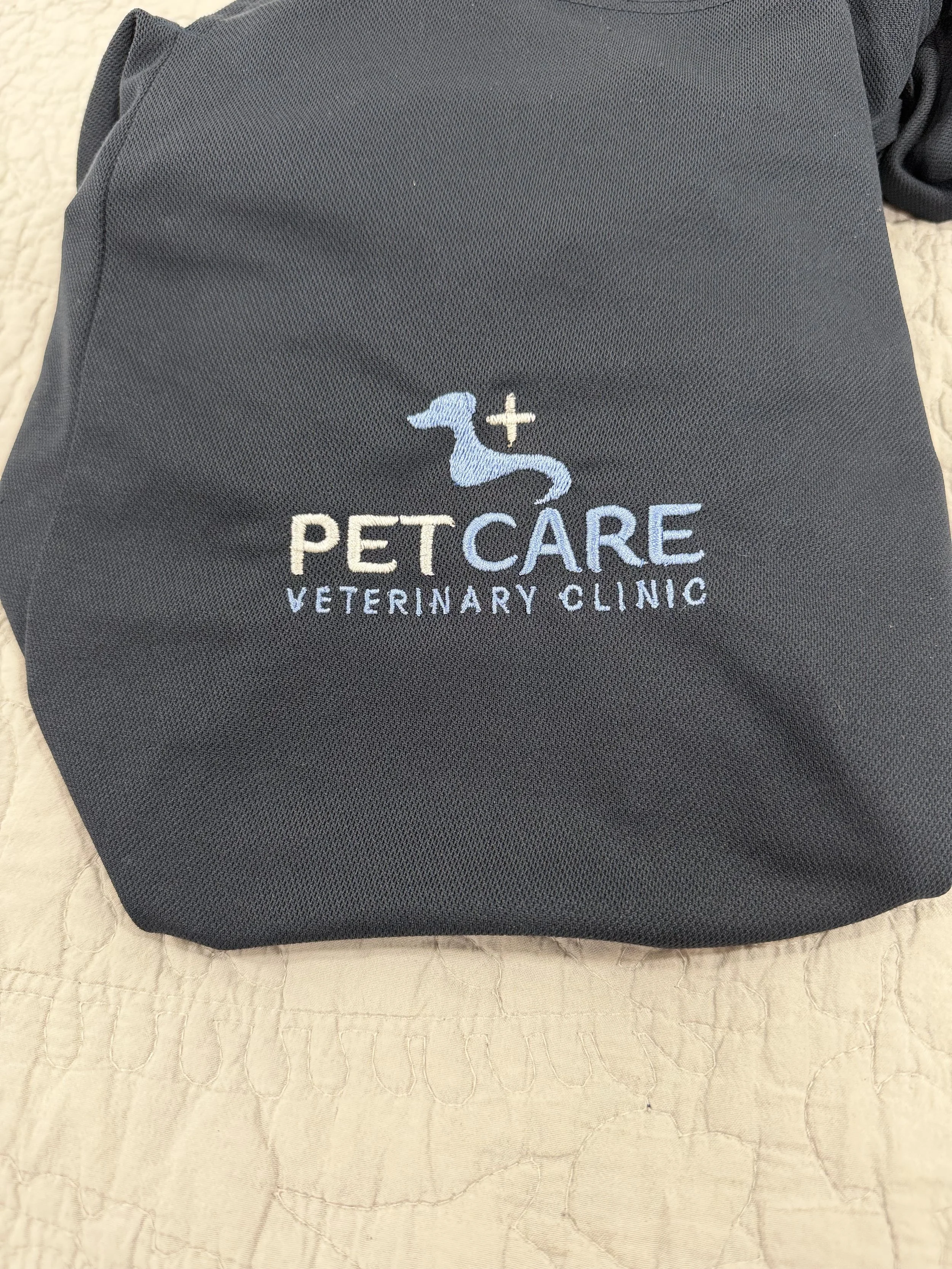 A custom embroidery example of a blue shirt with a PetCare Veterinary Clinic logo embroidered on it, featuring a dog silhouette, a cross, and the text 'PetCare Veterinary Clinic' on a quilted fabric surface.