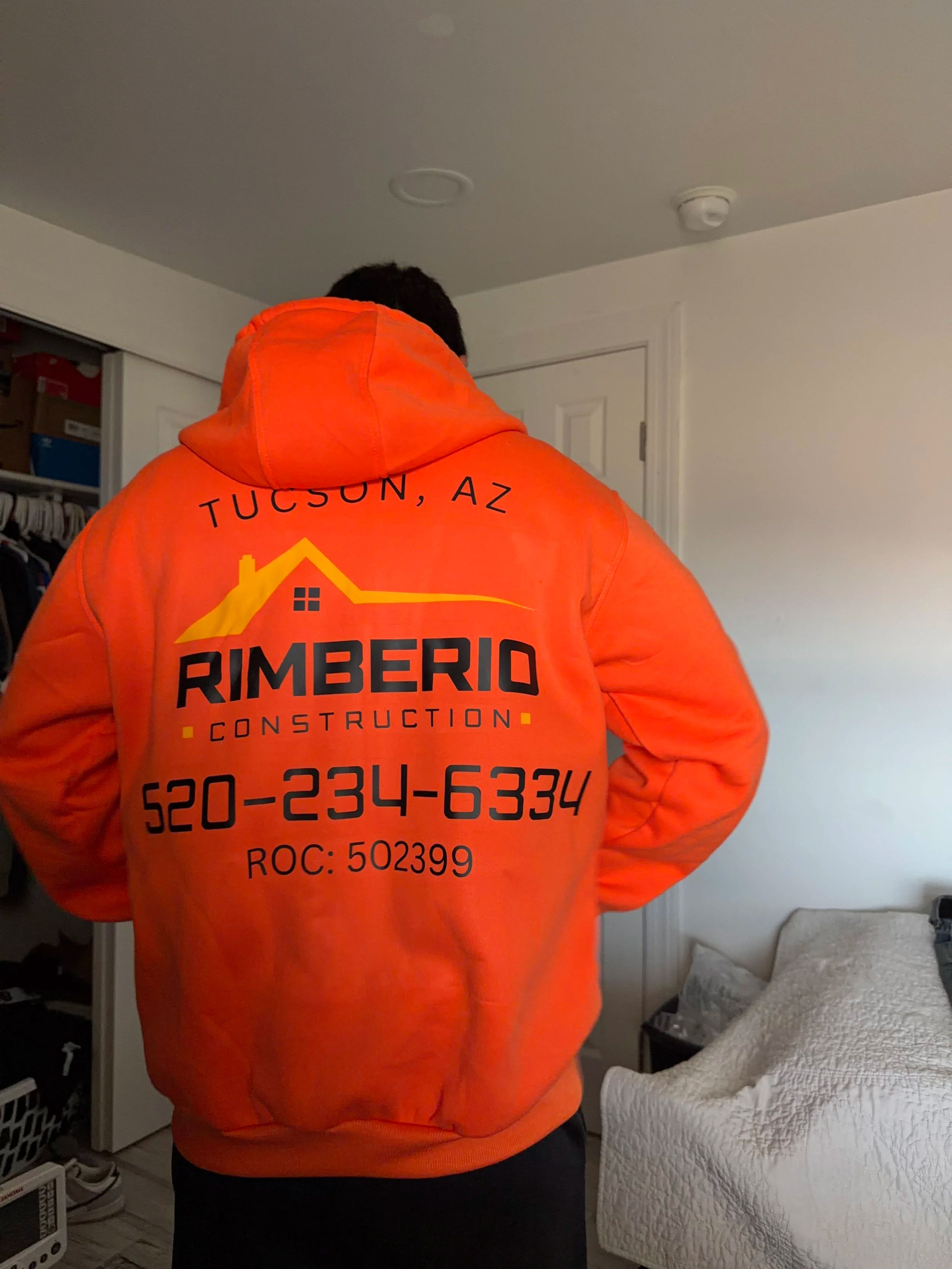 A custom direct to film print on a construction hoodie, back of a person wearing an orange hooded sweatshirt with company information for a remodeling construction business in Tucson, AZ, including a phone number and ROC license number.