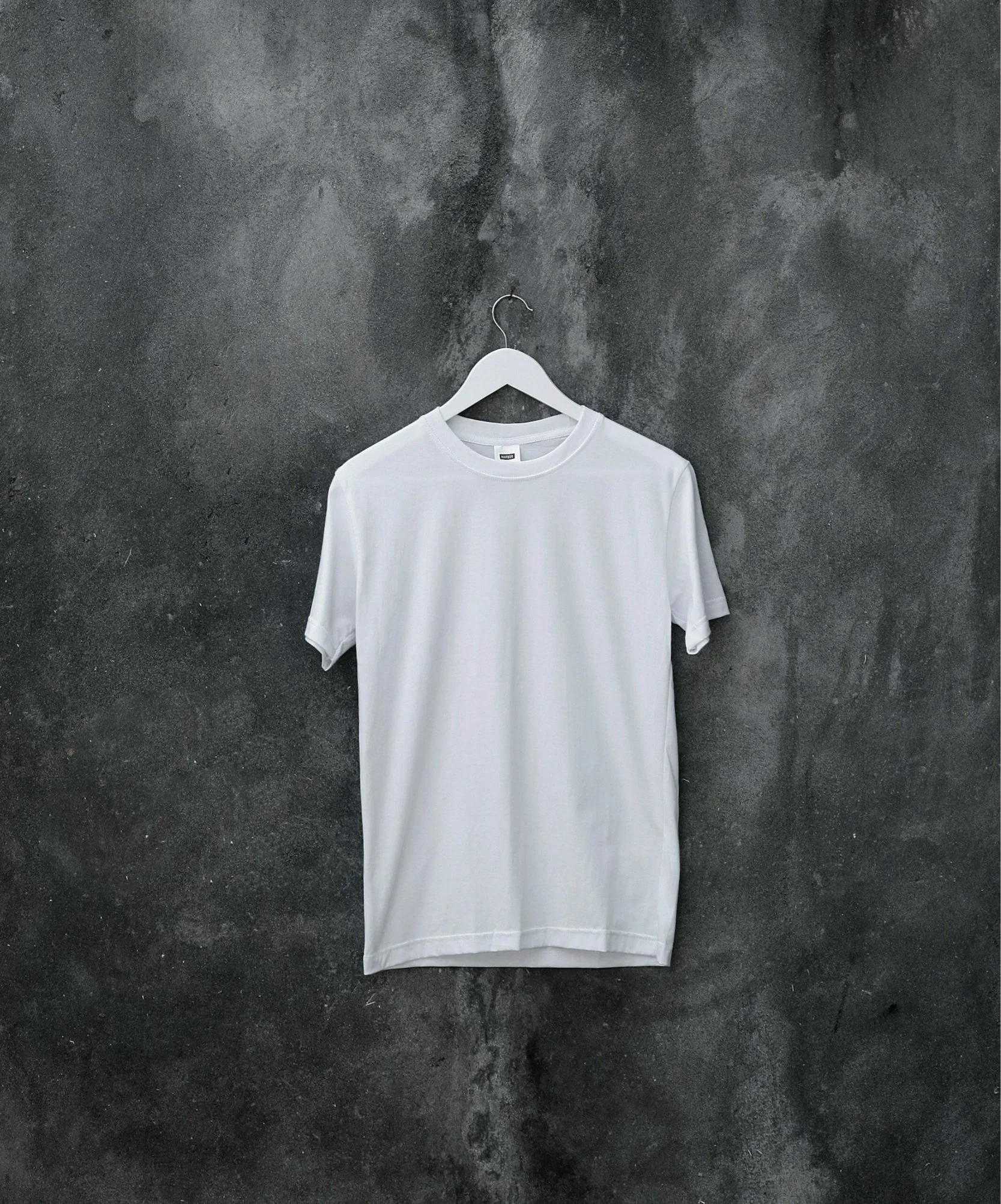 A plain white T-shirt hangs on a white hanger against a dark textured wall.