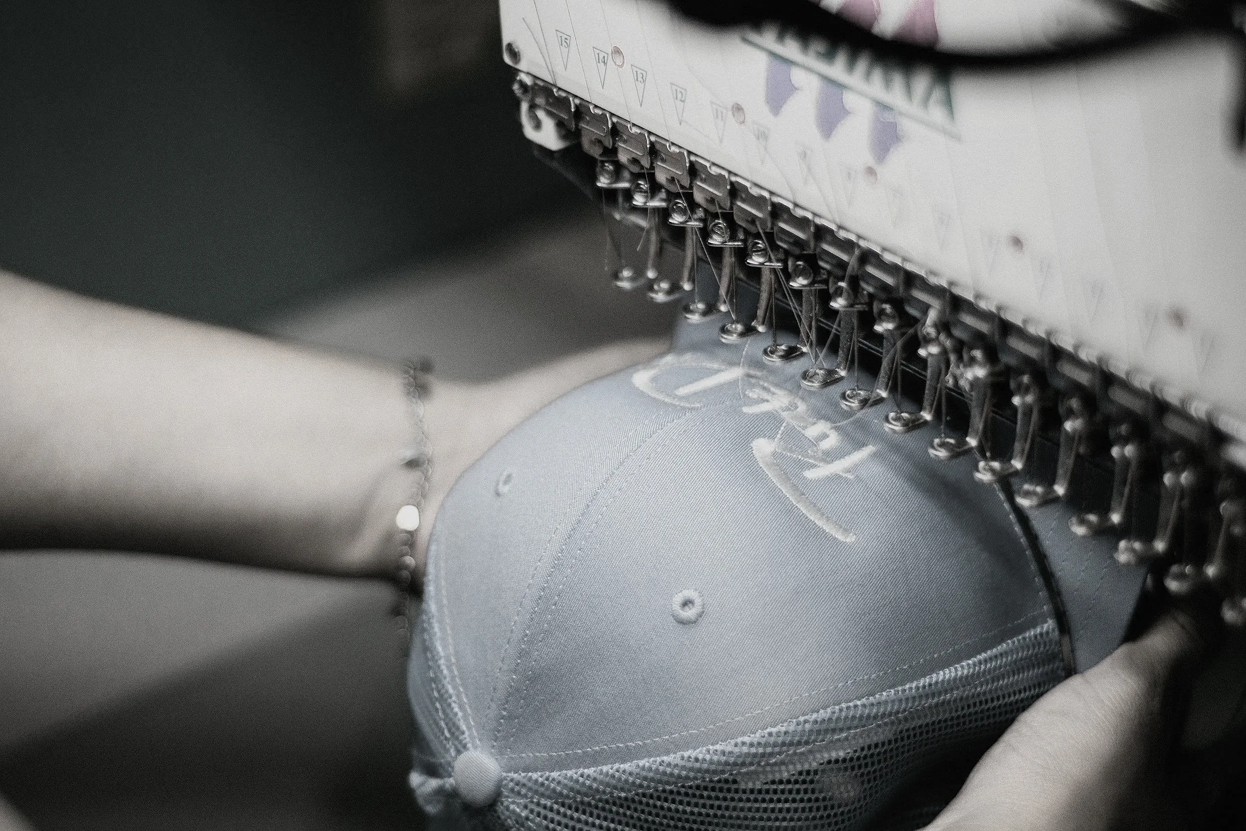 Close-up of a sewing machine stitching light gray fabric with embroidered letters, with a person's finger holding the fabric.