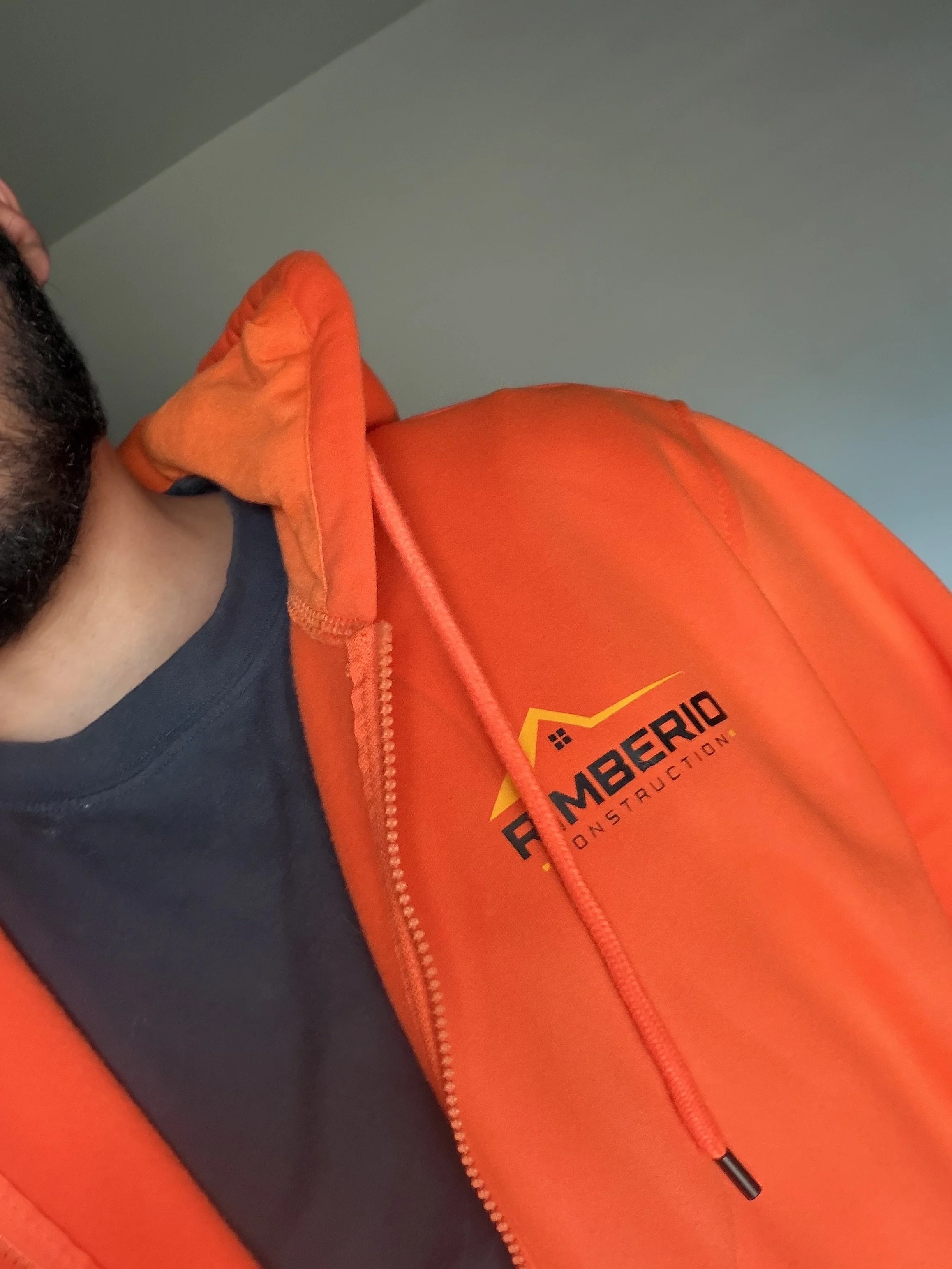Close-up of a person wearing a dark shirt and orange safety jacket with a logo that reads "FMBERIO CONSTRUCTION" with a stylized house icon, and a zipper sliding pull.