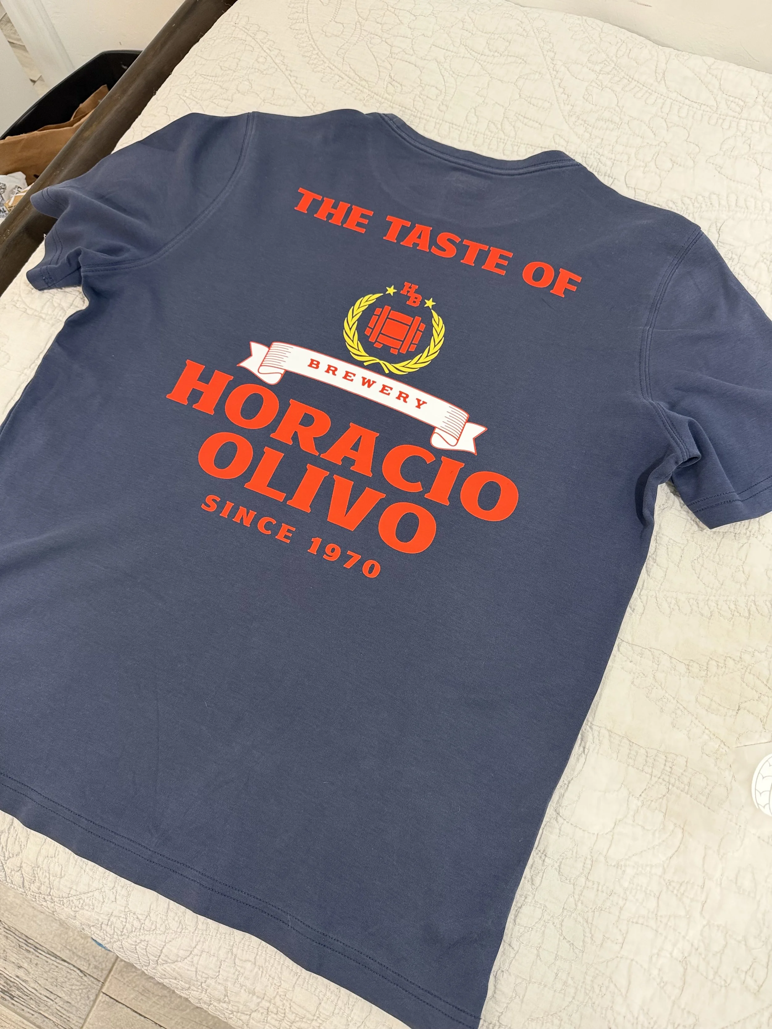 A blue t-shirt with custom dtf print of red, white, and yellow lettering and graphics that reads "The Taste of Horacio Olivo Brewery Since 1970" is laid flat on a quilted bedspread with a wooden headboard.