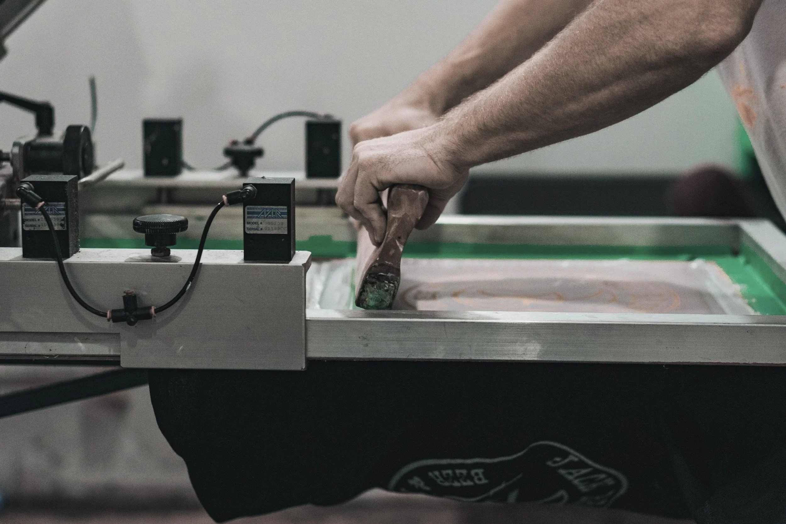 A person's arm is bending as they use a woodworking tool on a piece of wood, with a workbench and various electronic devices in the background.