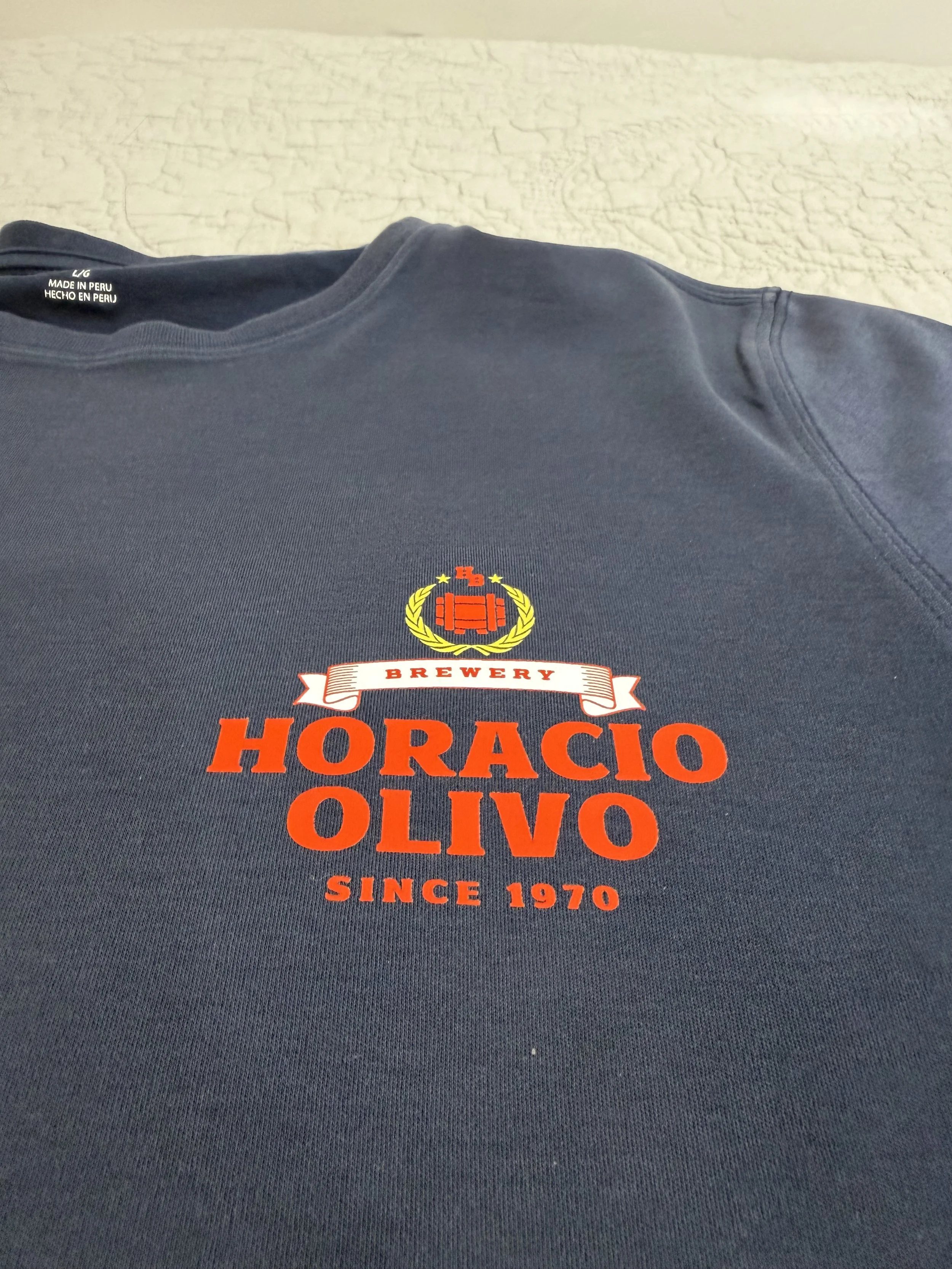 A custom example of a dtf shirt in navy blue T-shirt displays a logo with a red beer mug, surrounded by a yellow laurel wreath, and the words 'Brewery Horacio Olivo Since 1970' in orange and red text.