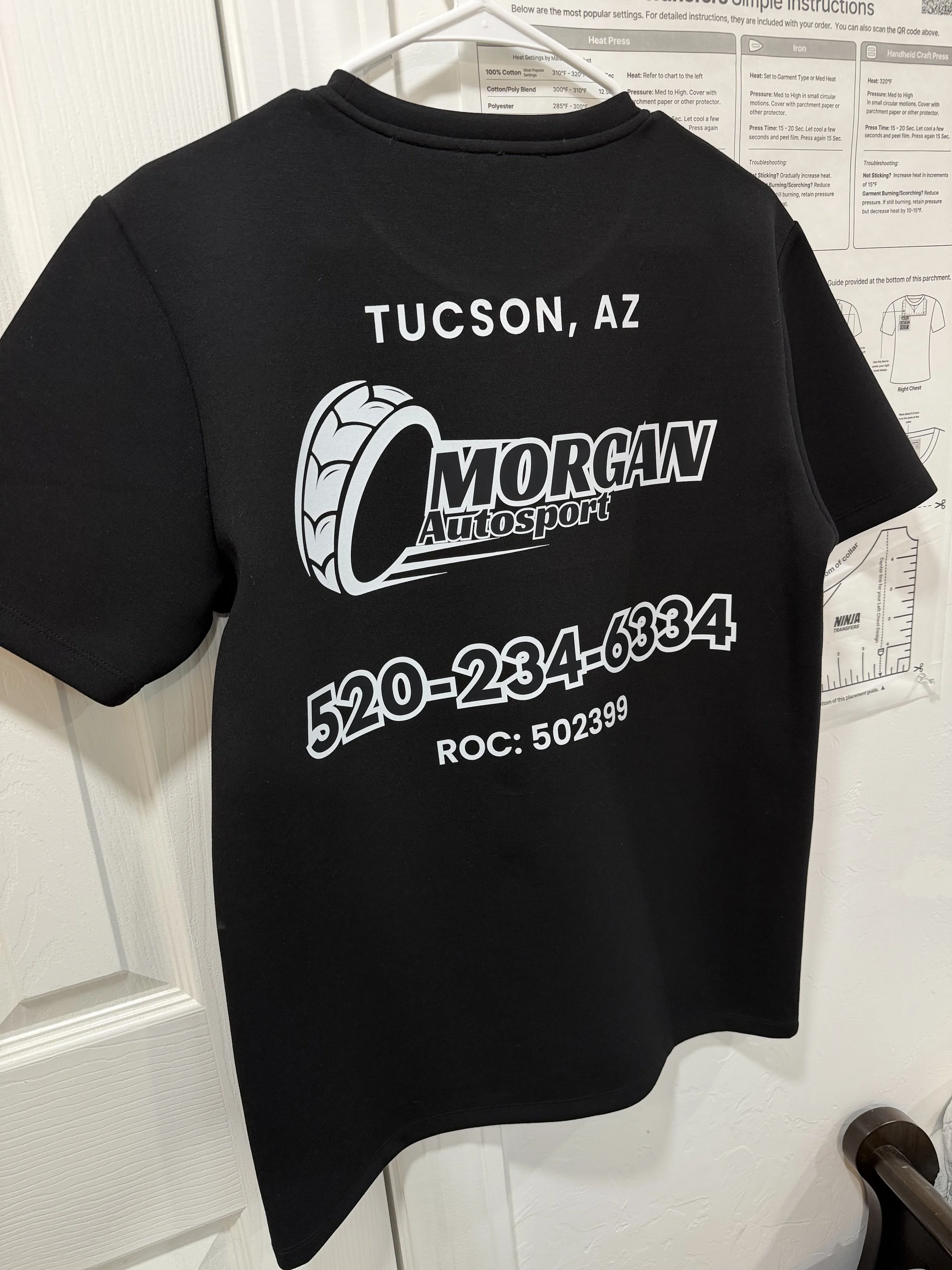 A custom DTF print black T-shirt hanging on a white hanger on a wall, with a graphic and text in white reading "TUCSON, AZ" and "Morgan Auto Sport" along with a stylized tire graphic and a phone number.