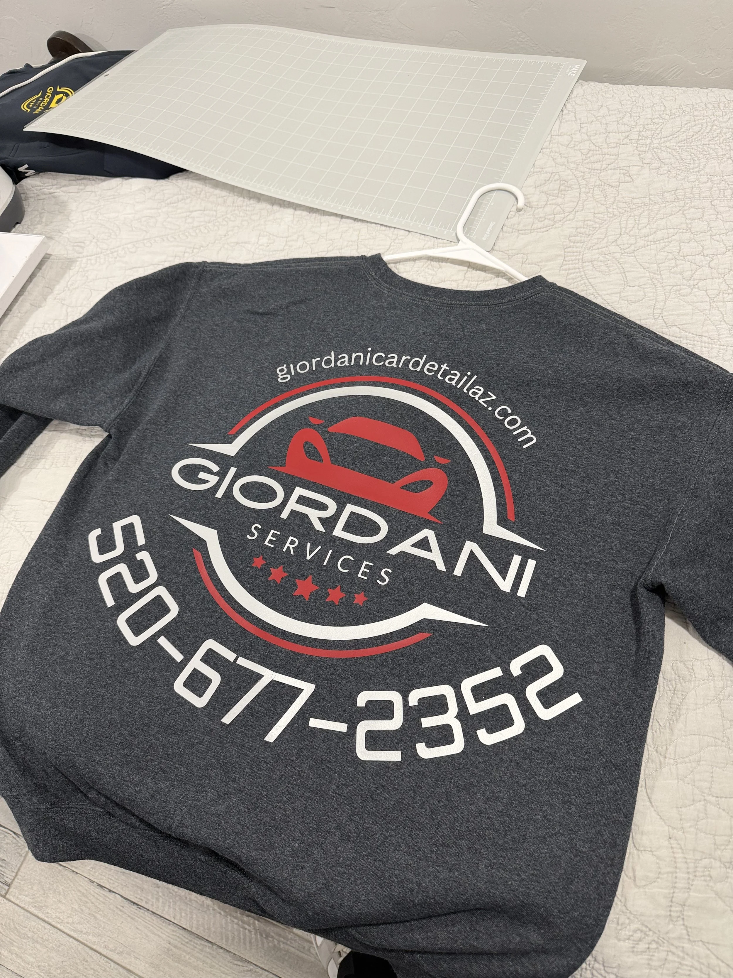 A custom print black T-shirt with a logo and website URL printed on it, placed on a bed next to a plastic cutting mat and a hanger, with a backpack and some items in the background.