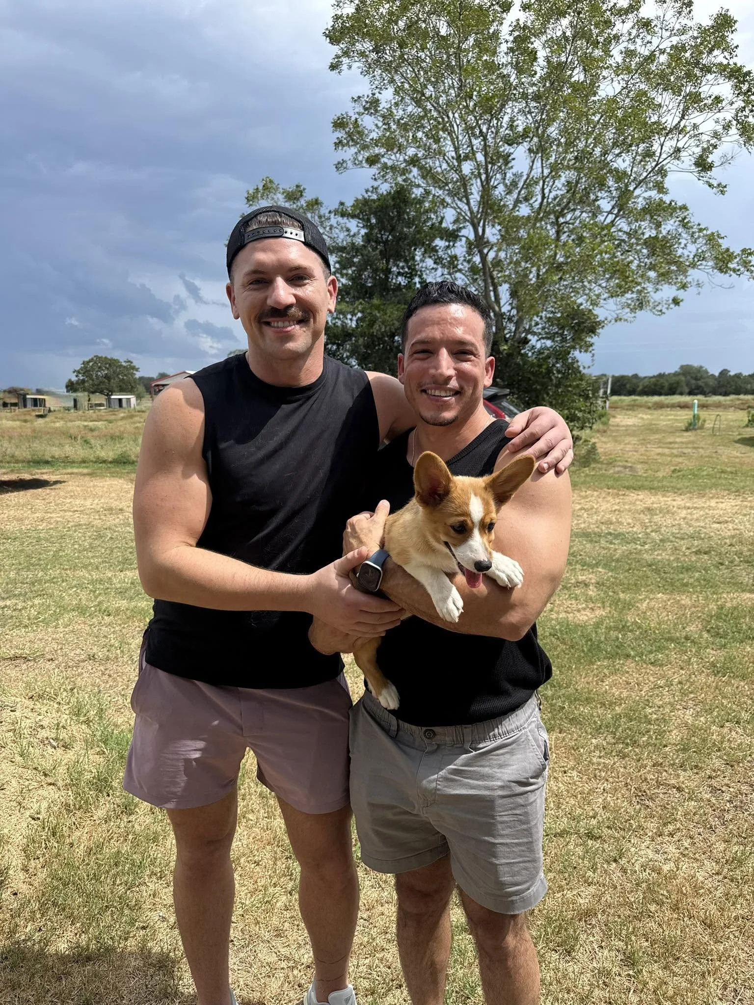 Two men standing outdoors in a grassy area, smiling, with a small dog in one of their arms. There are trees and a cloudy sky in the background.