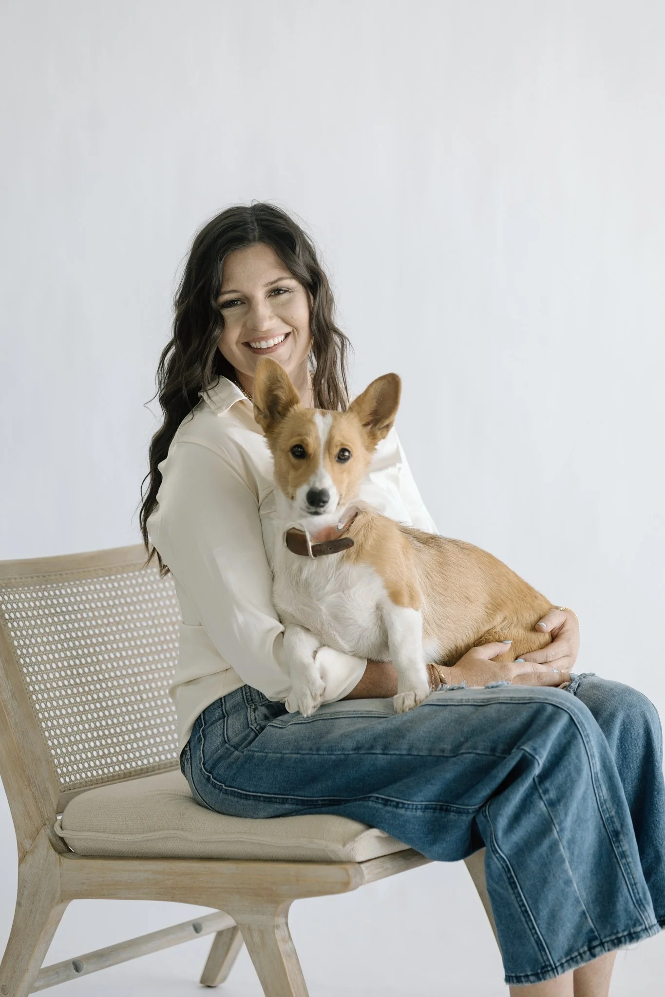 A woman sitting on a beige wooden chair with a woven backrest, smiling and holding a young tan and white dog on her lap, against a plain white background.