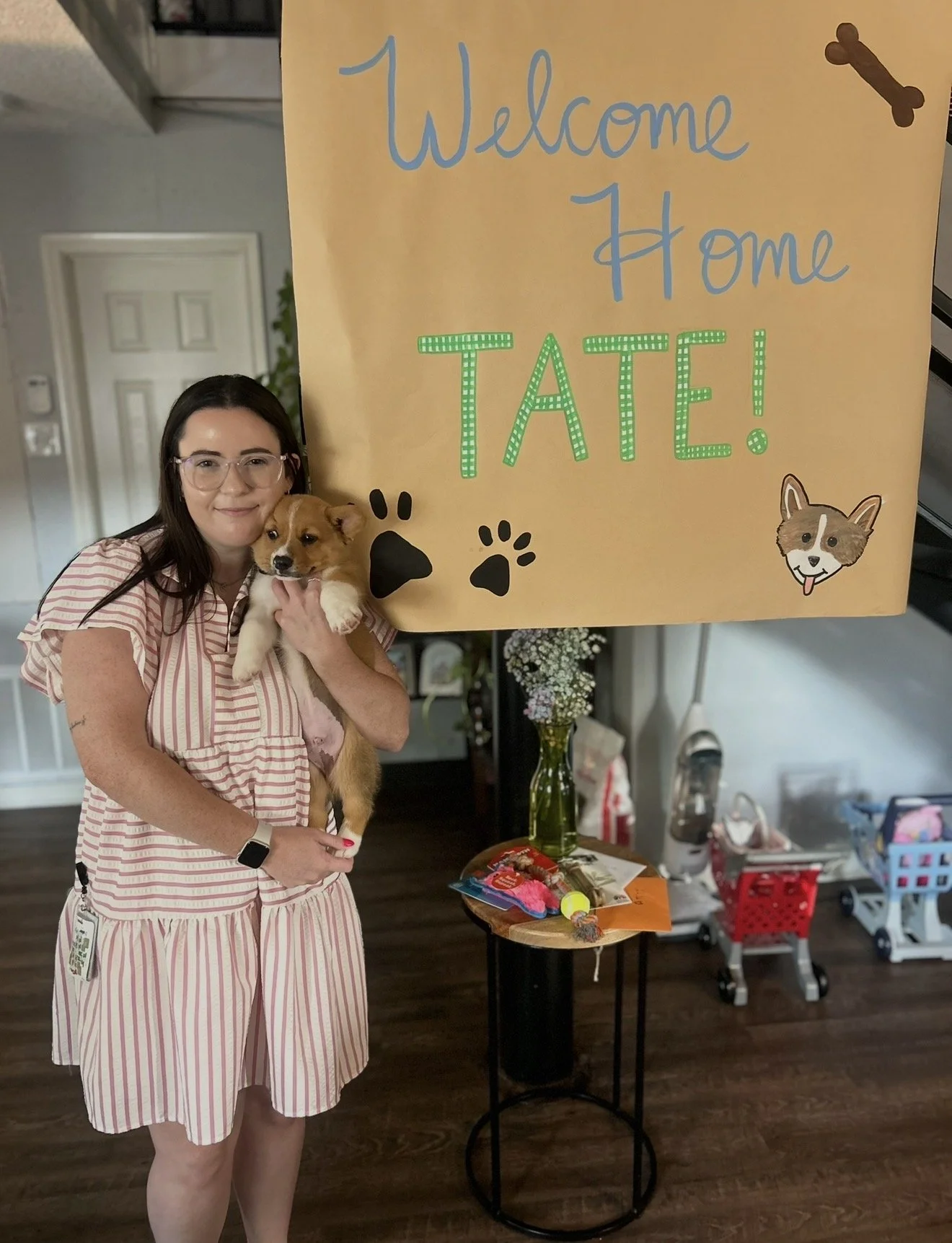 A woman in a pink and white striped dress holding a puppy in front of a large yellow sign that says "Welcome Home TATE!" in blue and green letters, decorated with dog paw prints, a dog bone, and a cartoon dog illustration. There is a table with dog toys and treats nearby, and a cluttered indoor space with toys and household items in the background.