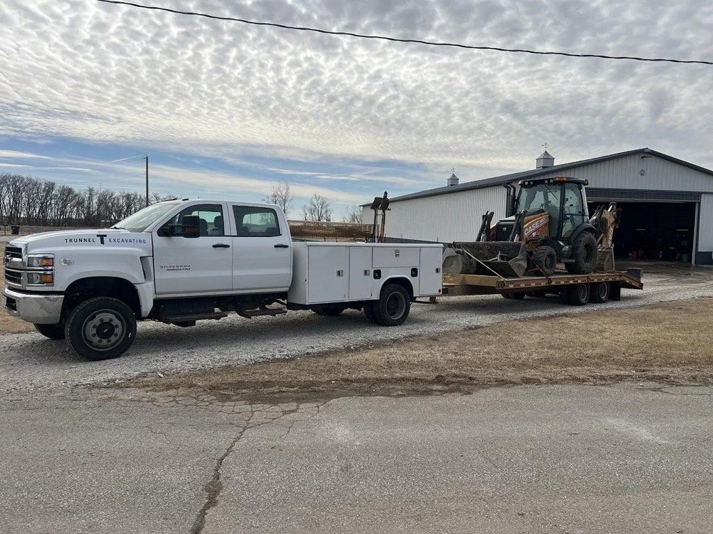 A white utility truck with a utility bed on the back is parked on a gravel road in front of a garage. The truck is carrying a small black and orange Case backhoe loader on a flatbed trailer. The background shows a rural area with leafless trees and an overcast sky.