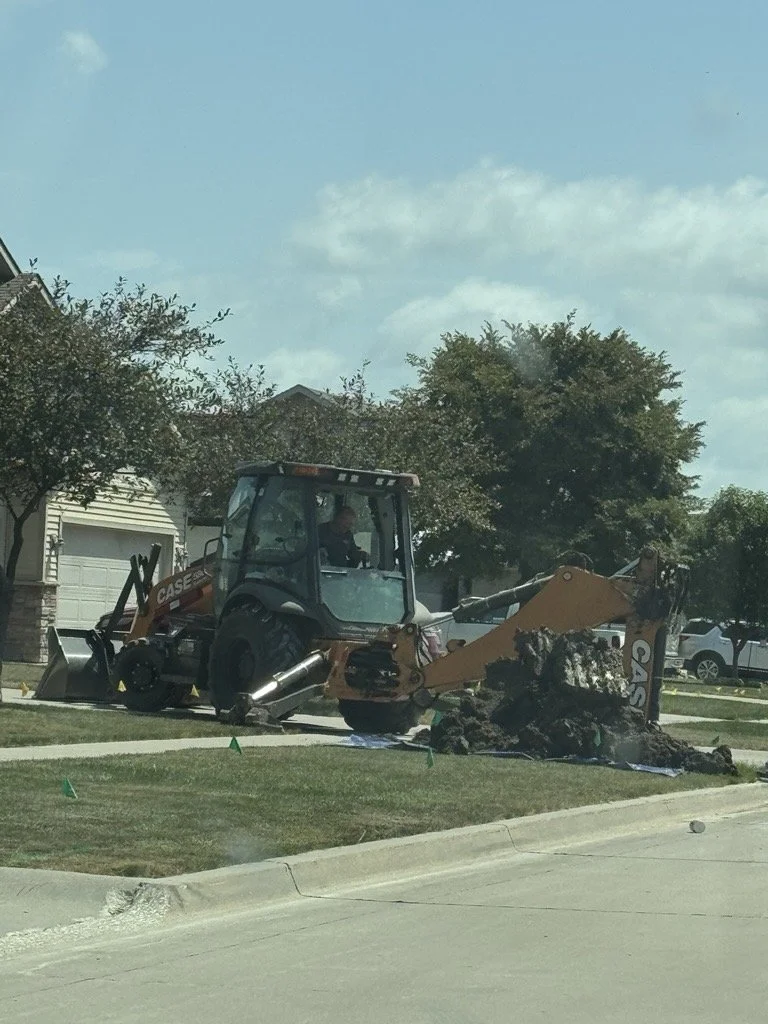 A construction vehicle, specifically a small excavator, is working on the front yard of a residential neighborhood, digging a hole near the sidewalk.